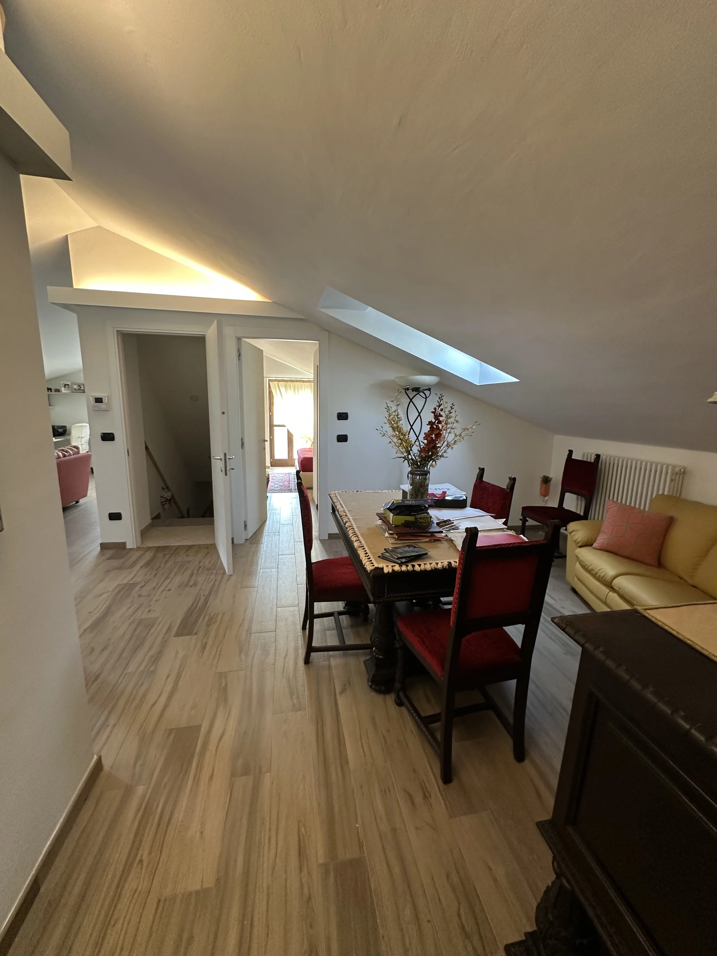 Interno di una casa con soffitto inclinato, tavolo con libri e fiori, divano giallo e sedie rosse.