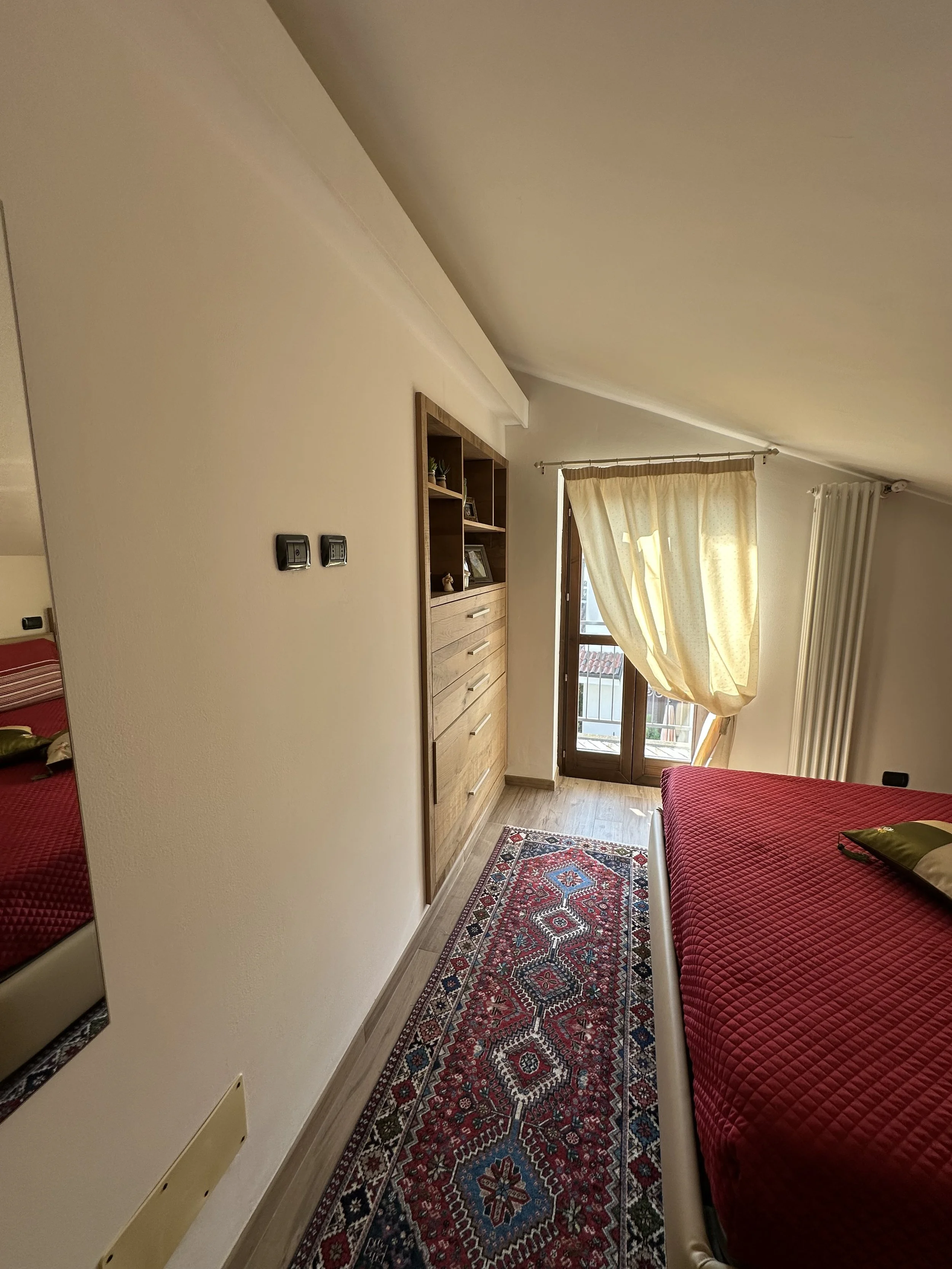 Camera da letto con letto con copriletto rosso, armadio in legno, tappeto persiano, finestra con tende gialle e porta finestra che dà su un balcone.