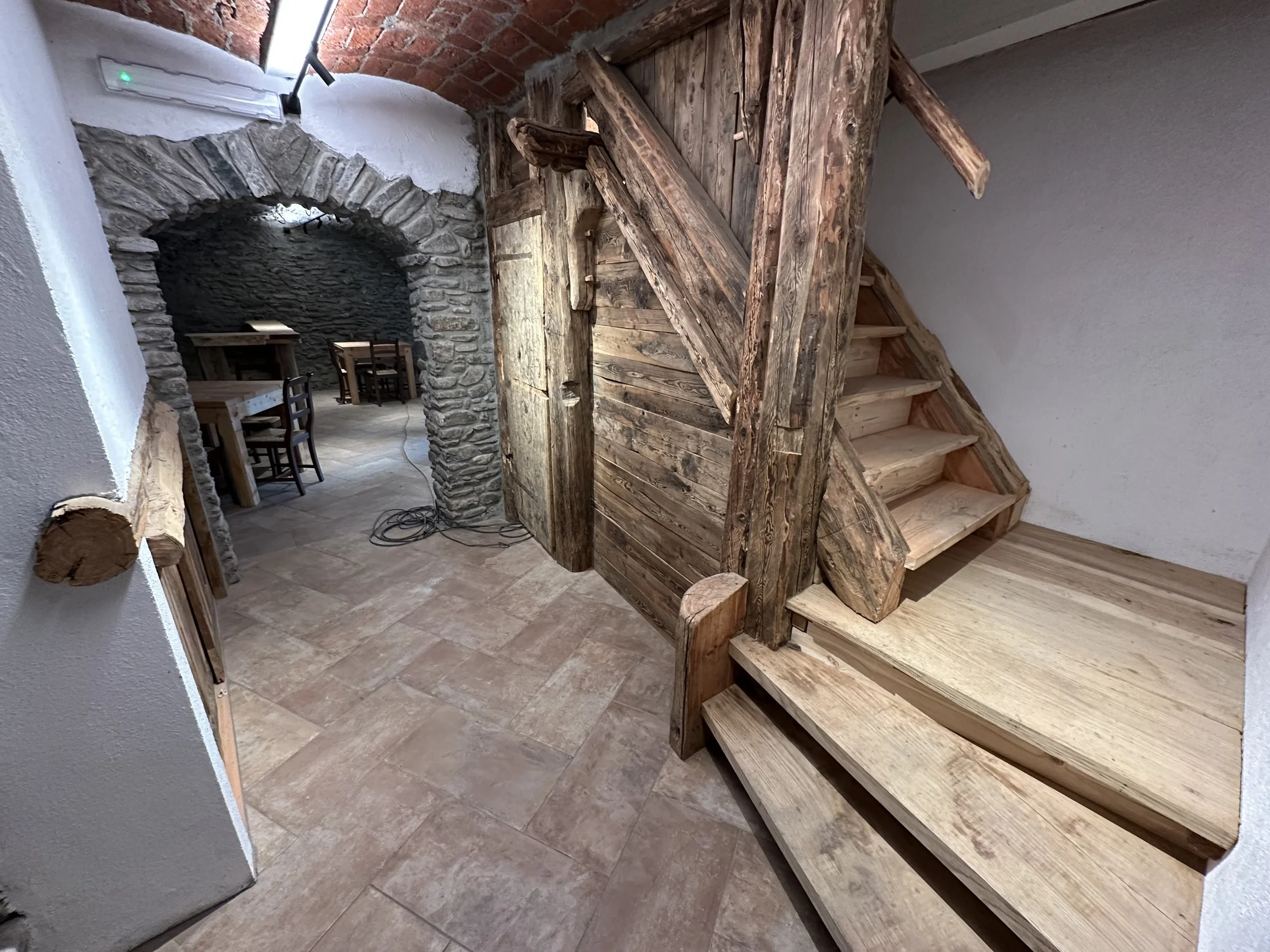 Interno di una stanza con scala in legno, pareti in pietra e mattoni, e arredi in stile rustico.