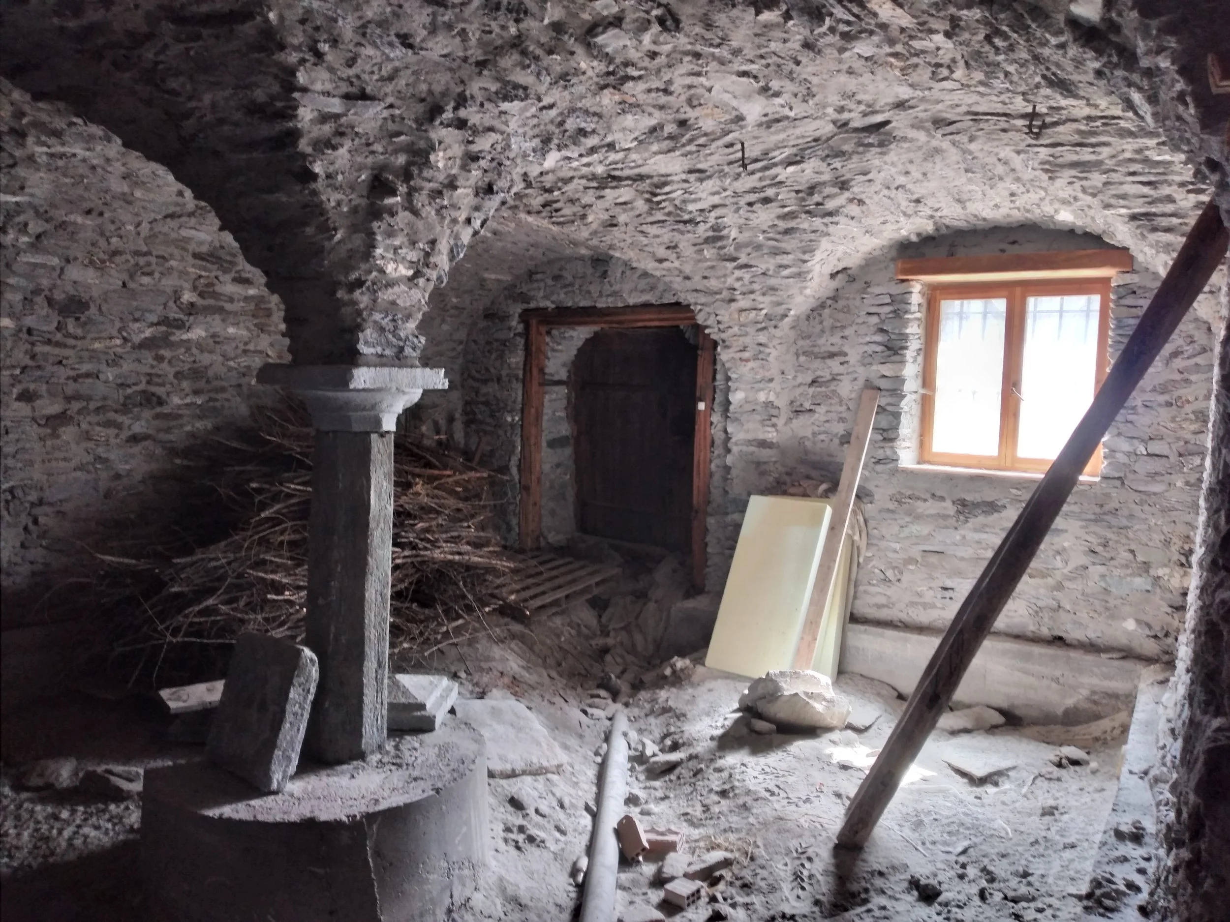 Interno di una stanza in costruzione con pareti di pietra, finestra con cornice in legno, strumenti da costruzione e materiali vari sul pavimento.