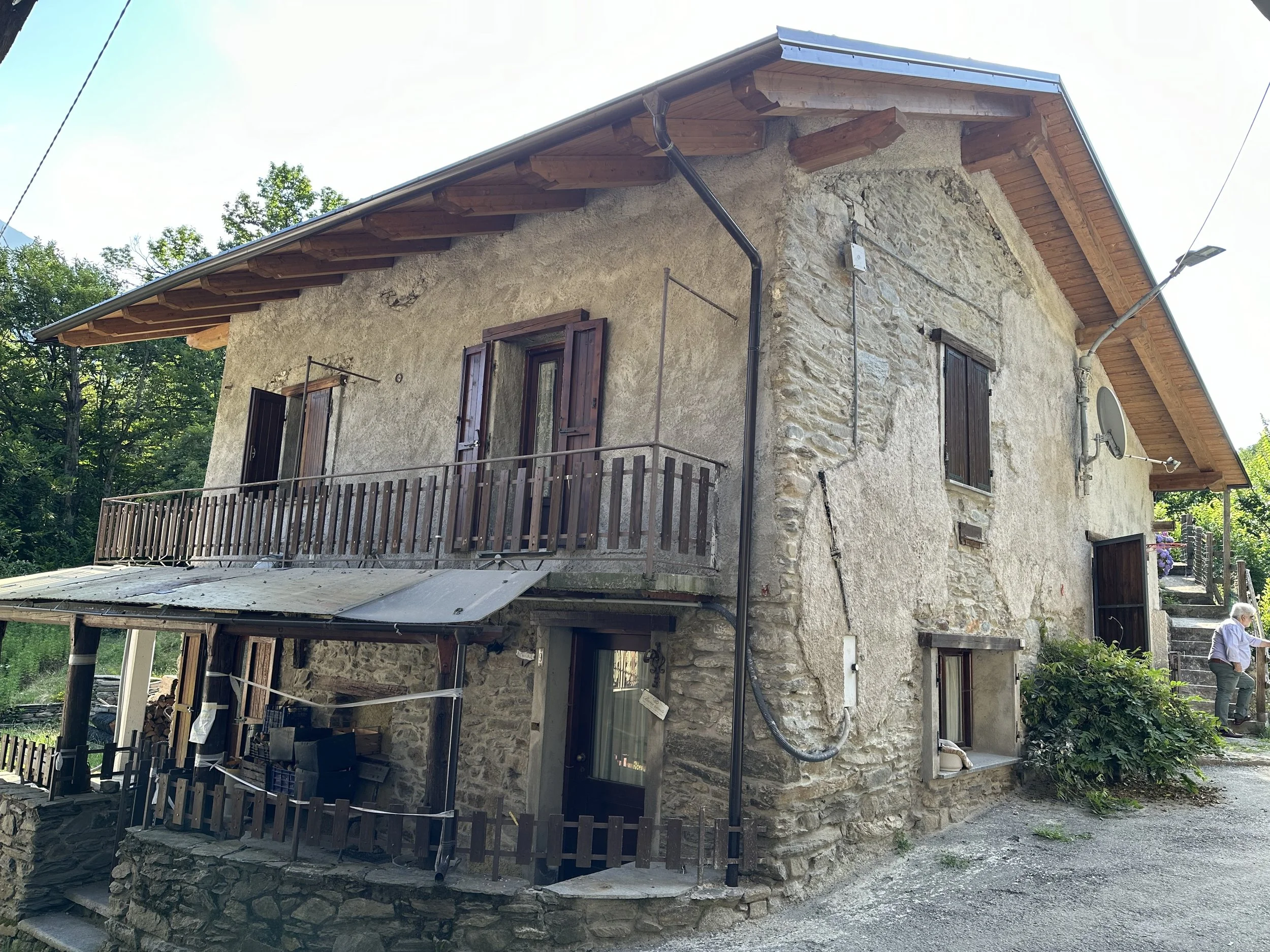 Casa di montagna in pietra con balcone e finestre con persiane di legno, circondata da alberi e vegetazione.