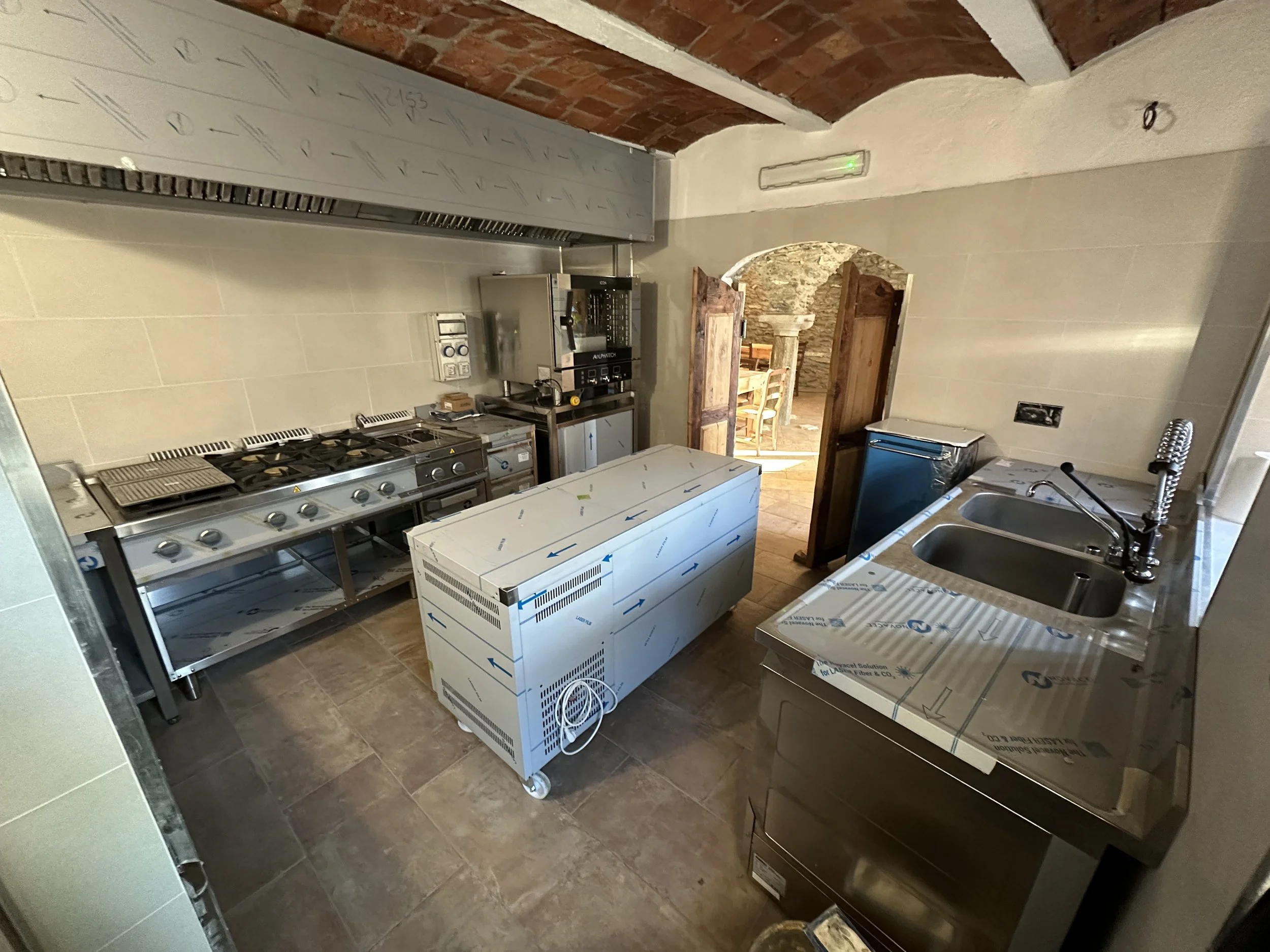 Cucina in fase di ristrutturazione con mobili nuovi e ripiani in acciaio; porta aperta che conduce a una stanza con tavolo e sedie in pietra