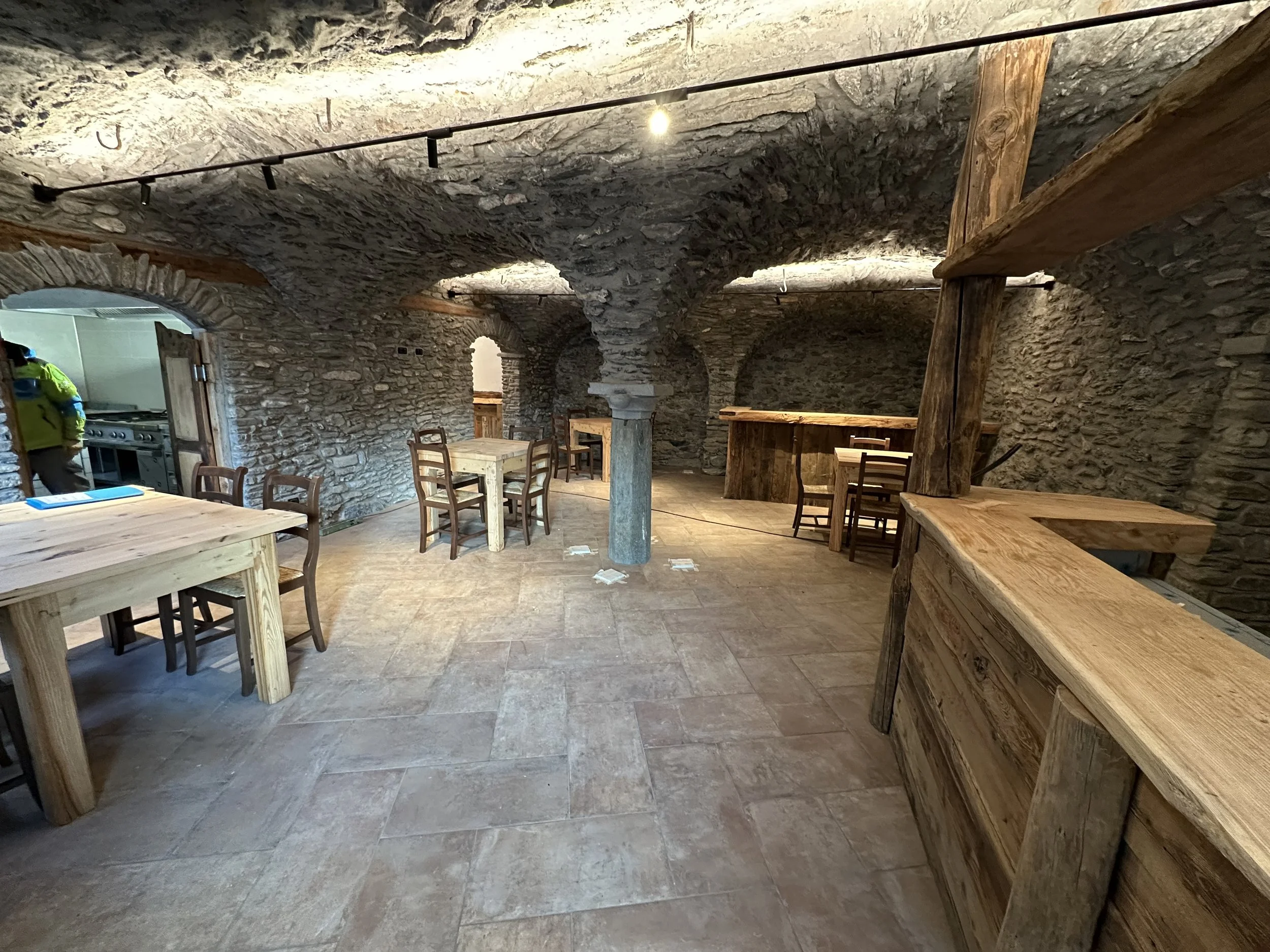 Interno di una cantina con muri in pietra, tavoli e sedie in legno, arredi rustici e archi in pietra, illuminazione calda.