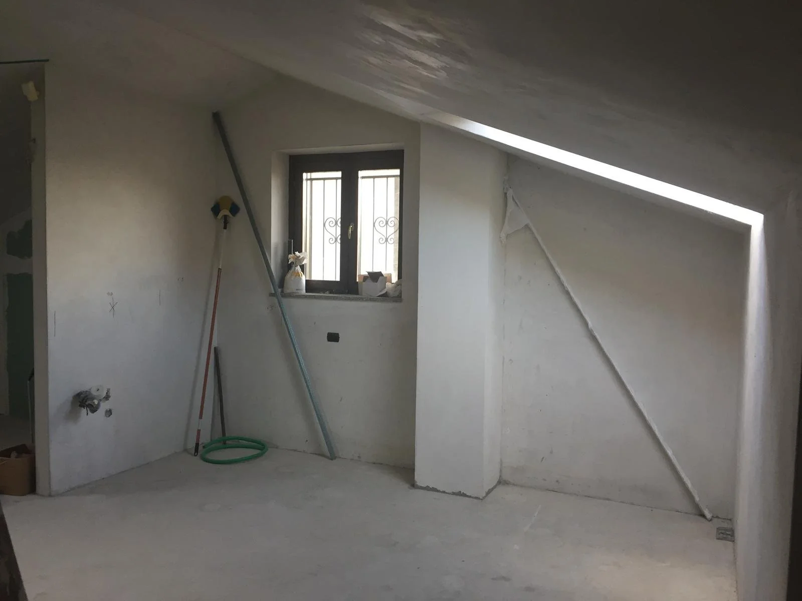 Interno di una stanza sottotetto in costruzione, senza finiture, con una finestra e alcuni attrezzi da lavoro appoggiati alla parete.
