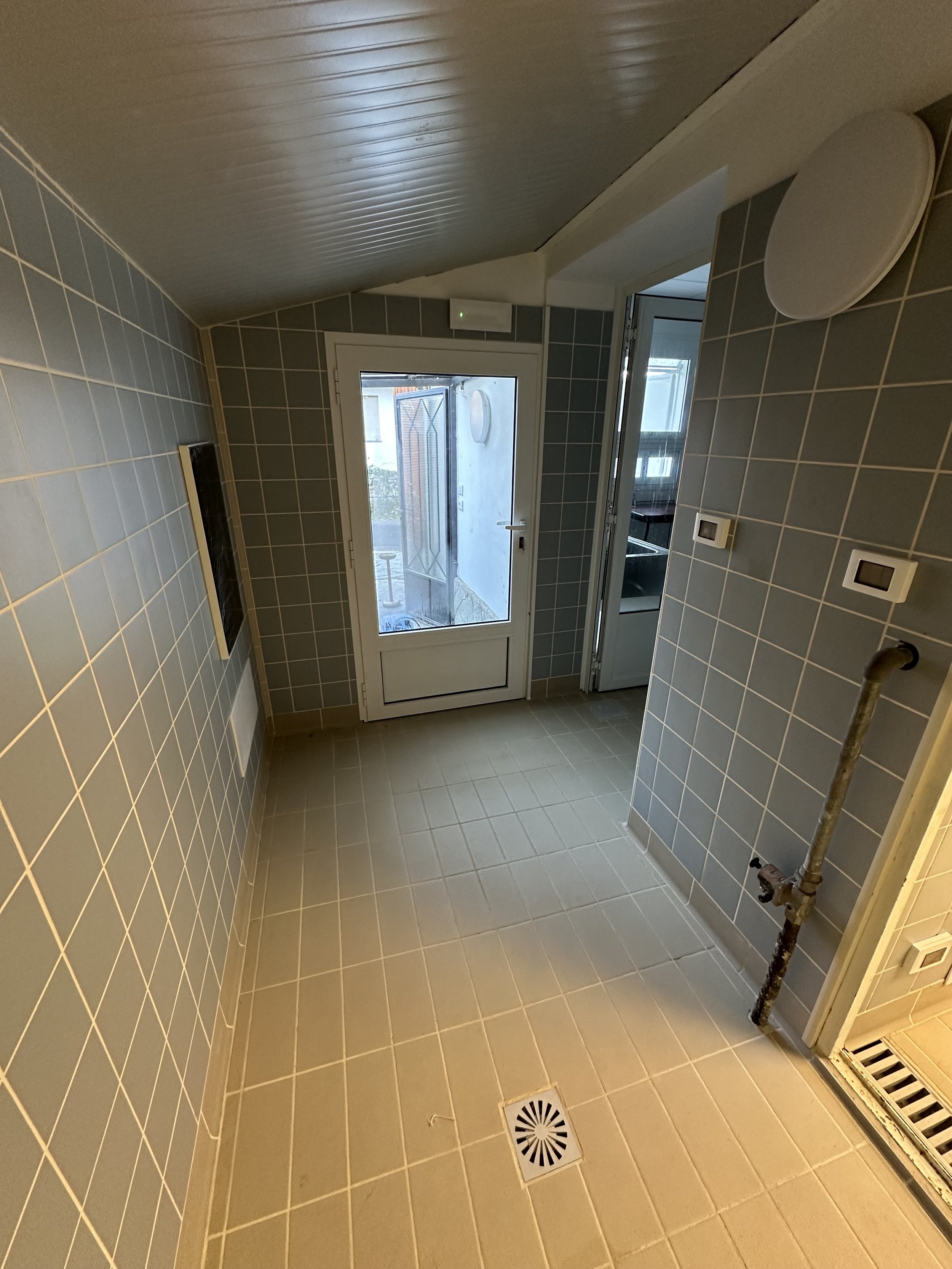 Interno di una stanza con pareti e pavimenti rivestiti di piastrelle, porta finestra che conduce all'esterno, e alcuni elementi come un pannello, un termostato e un tubo con valvola.