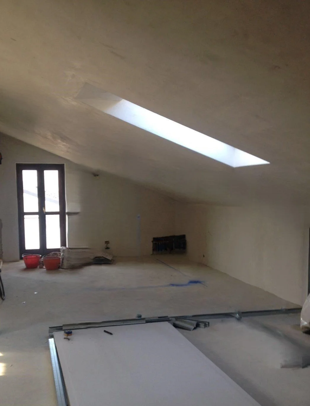 Interno di una stanza sotto ristrutturazione con pareti e soffitto in fase di finitura, una finestra e una grande apertura sul soffitto che permette l'ingresso di luce naturale.