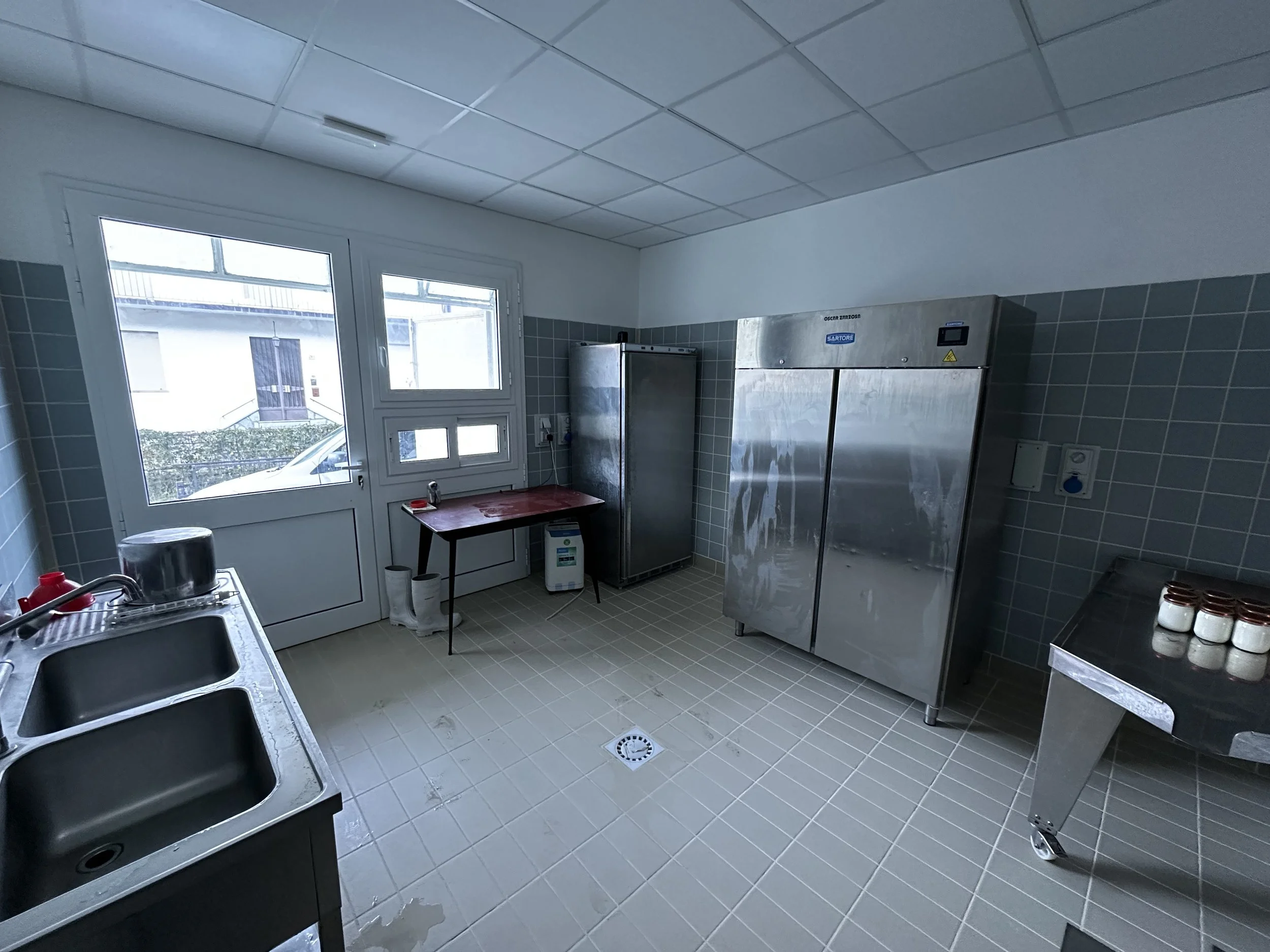 Cucina o locale di lavaggio con lavandino, frigorifero, armadietti in acciaio, tavolo con alcune bottiglie, piastrelle sul pavimento e sulle pareti, finestra con vista all'esterno.