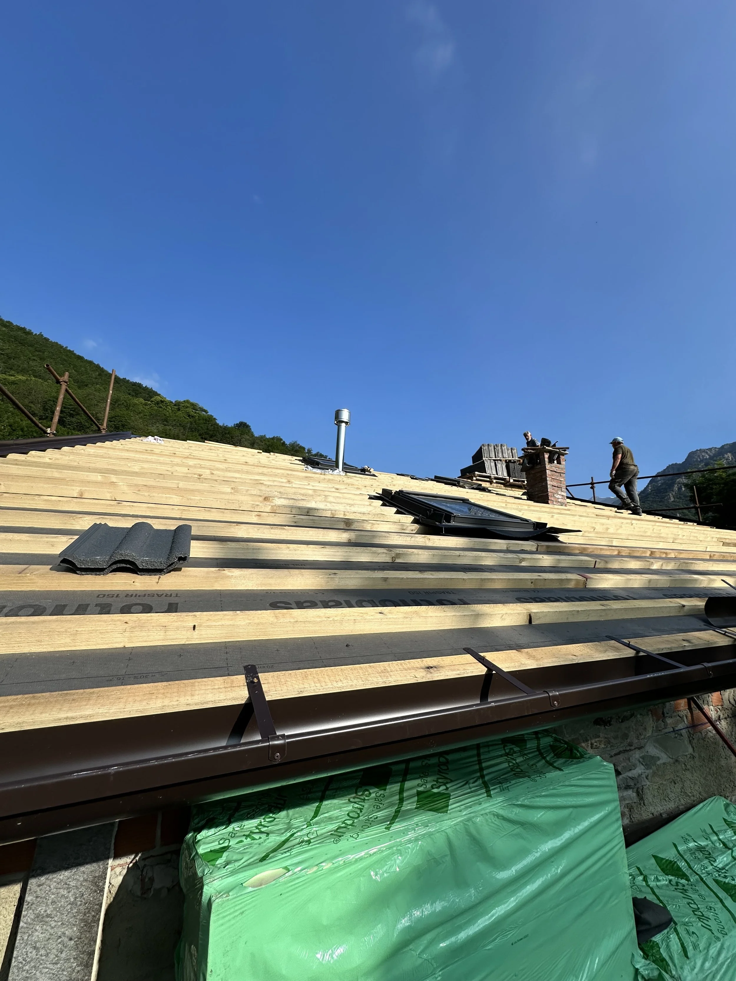 Lavori edilizi sul tetto di una casa in costruzione, con attrezzi e materiali, due operai e un cielo blu.