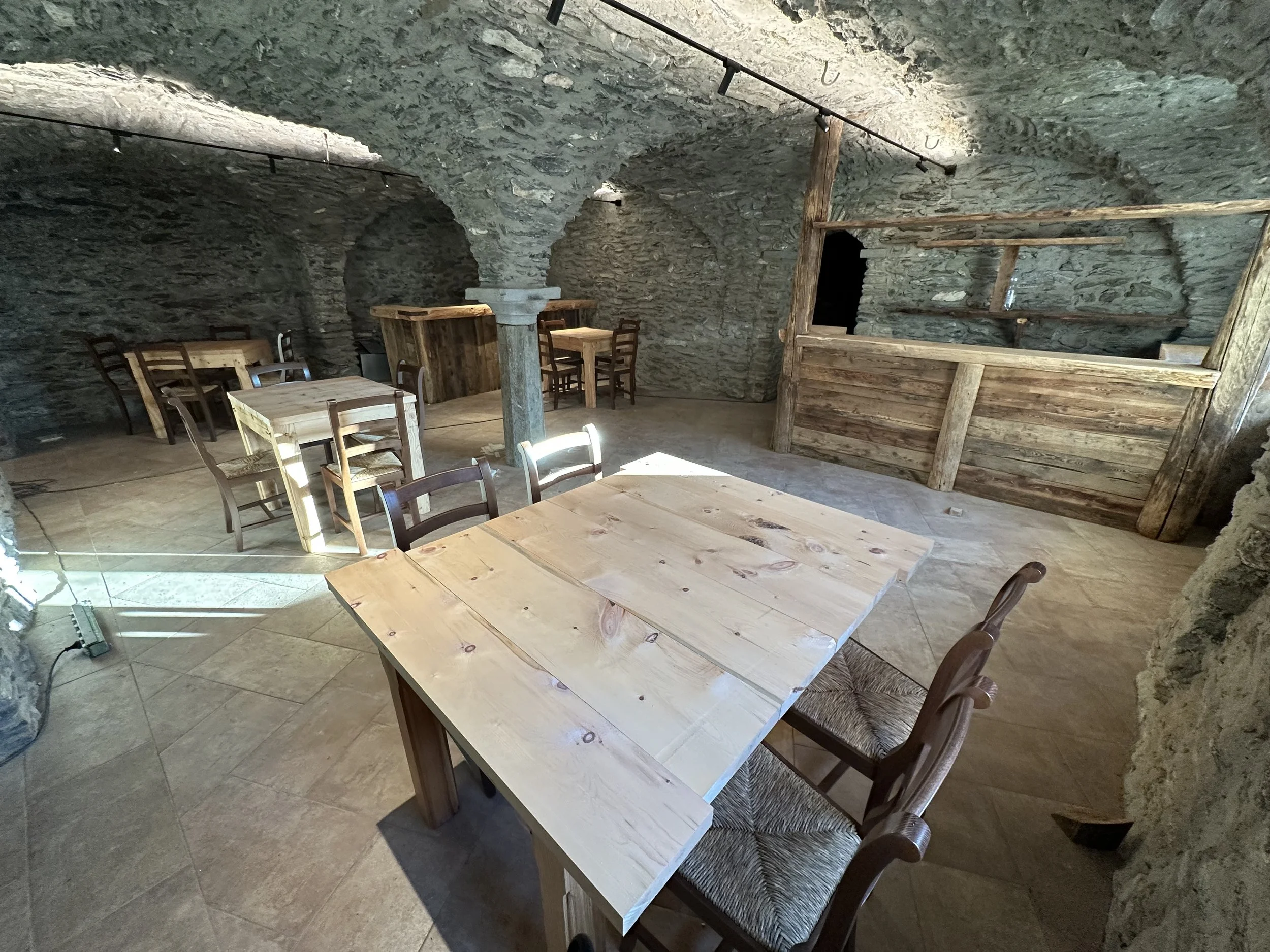 Interno di un ristorante sotterraneo con pareti di pietra, tavoli di legno e sedie in legno, illuminazione naturale e strutture in legno.