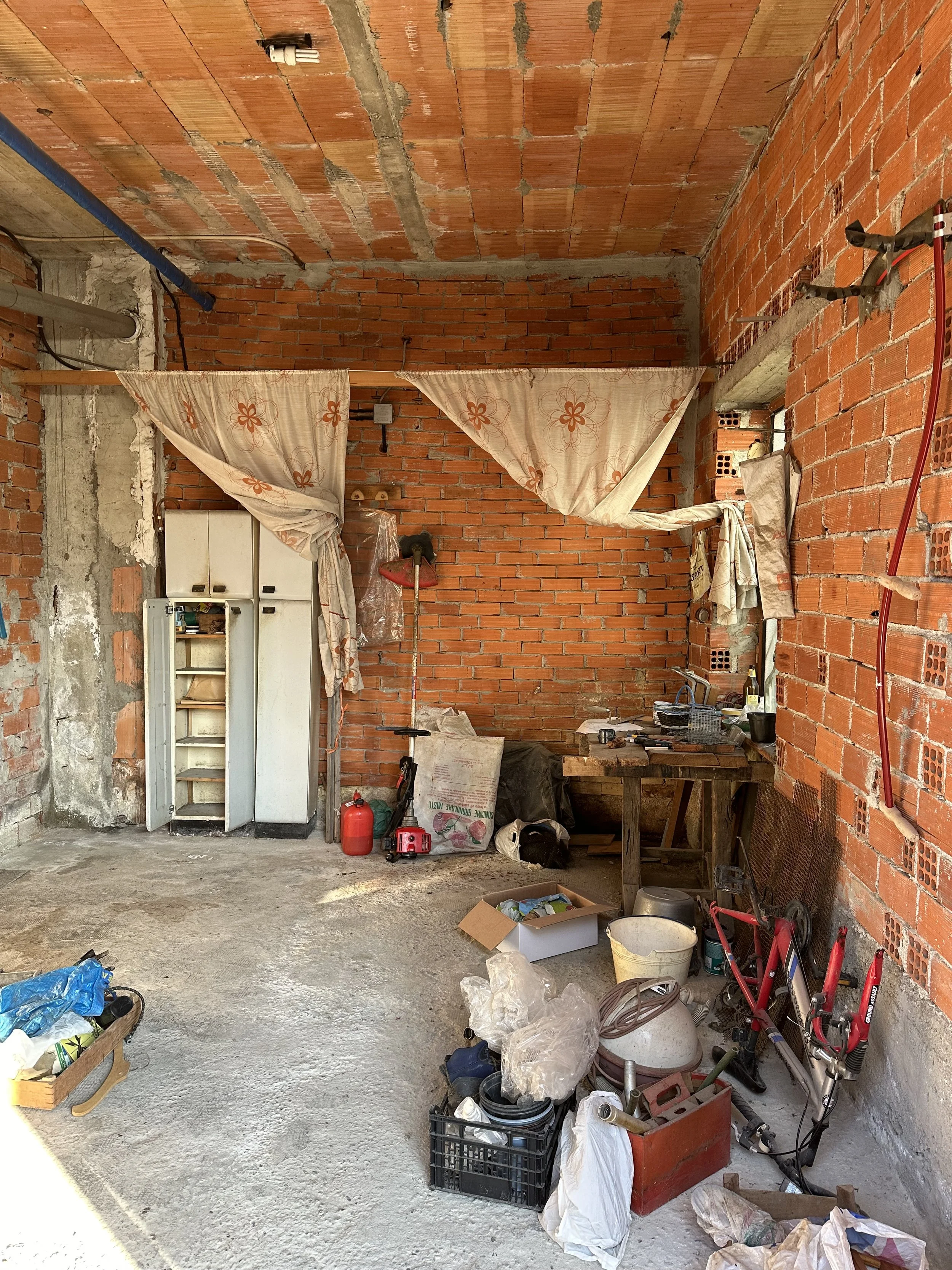Interno di un garage in costruzione con pareti di mattoni rossi, scatole di utensili, biciclette e materiali vari lasciati a terra.