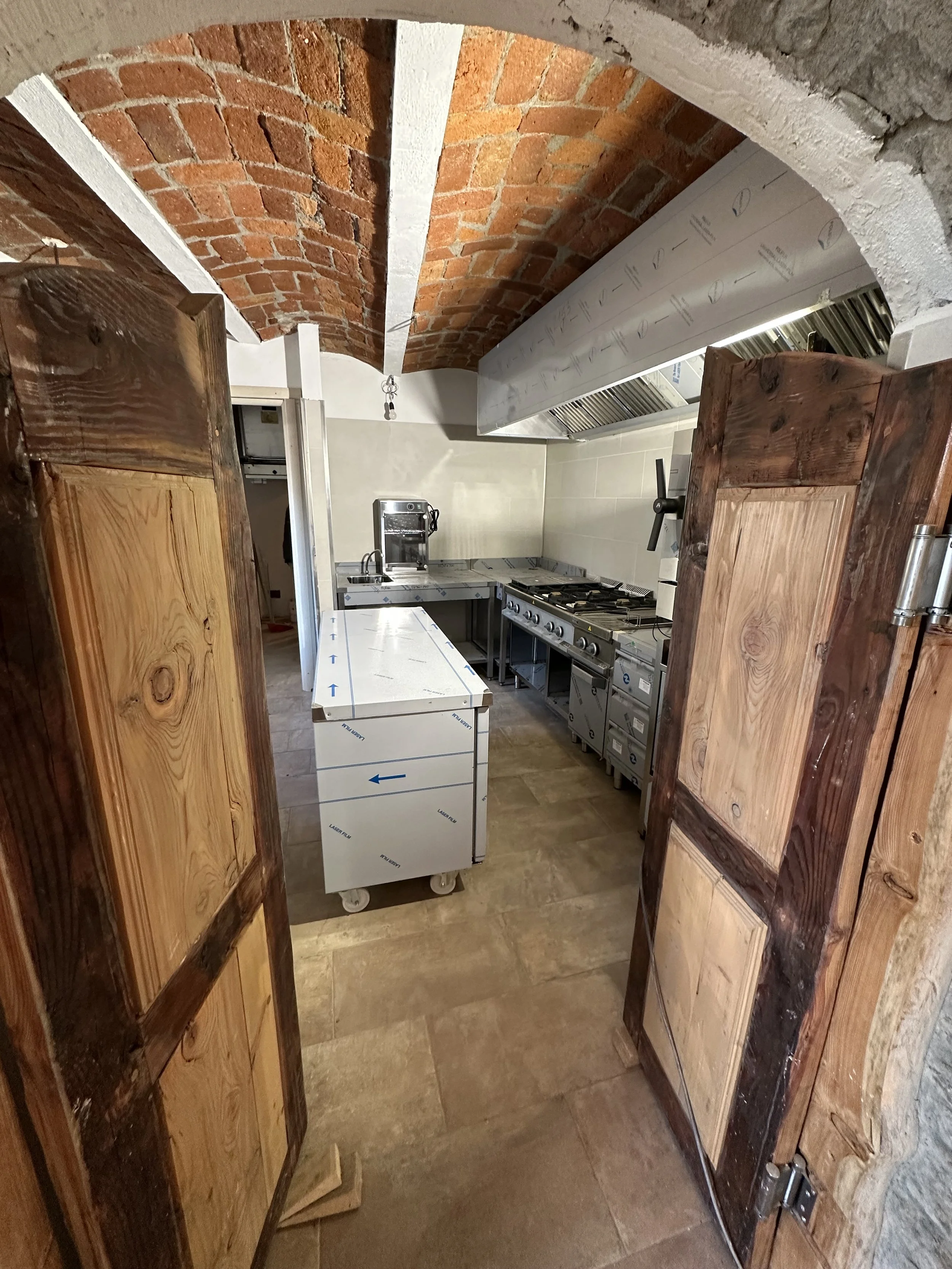 Cucina professionale in fase di ristrutturazione, con pareti in mattoni e attrezzature inox, vista attraverso due ante di legno.
