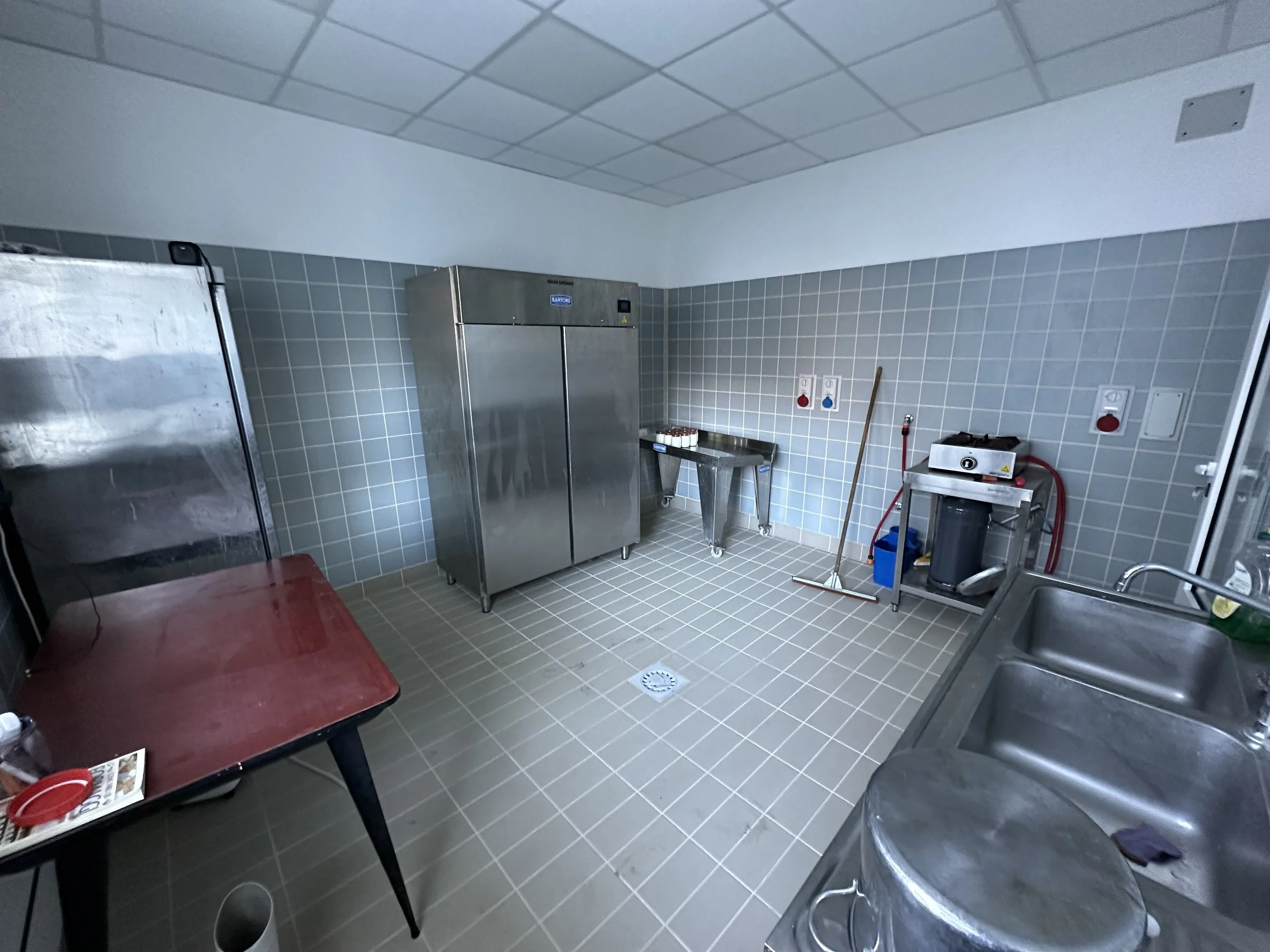 Stanza di cucina o laboratorio con sink, frigorifero e tavolo di lavoro in acciaio, pareti e pavimenti rivestiti di piastrelle grigie.