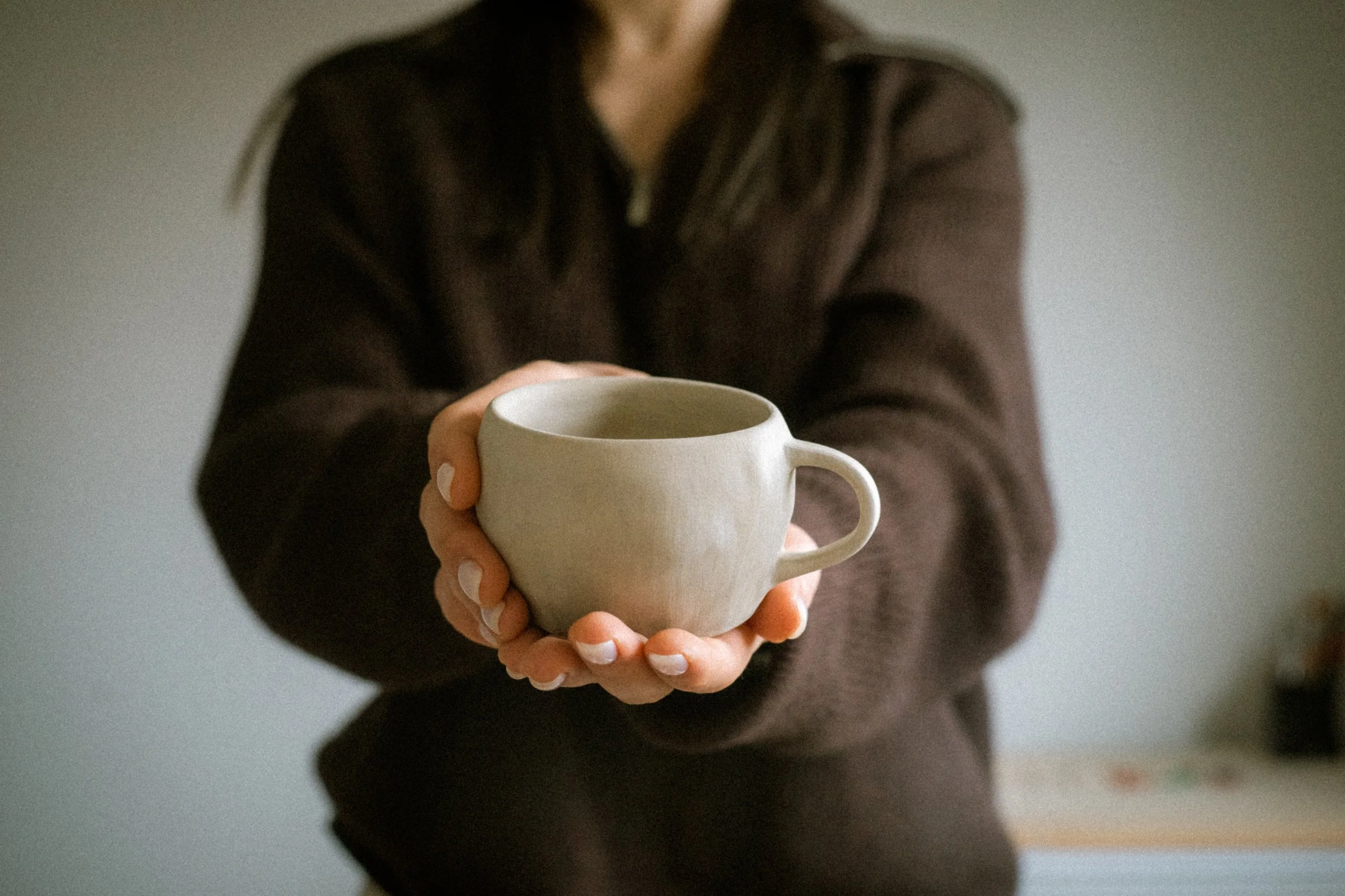 Person in a brown sweater holding a bone-dry ceramic mug with both hands, extending it toward the camera.