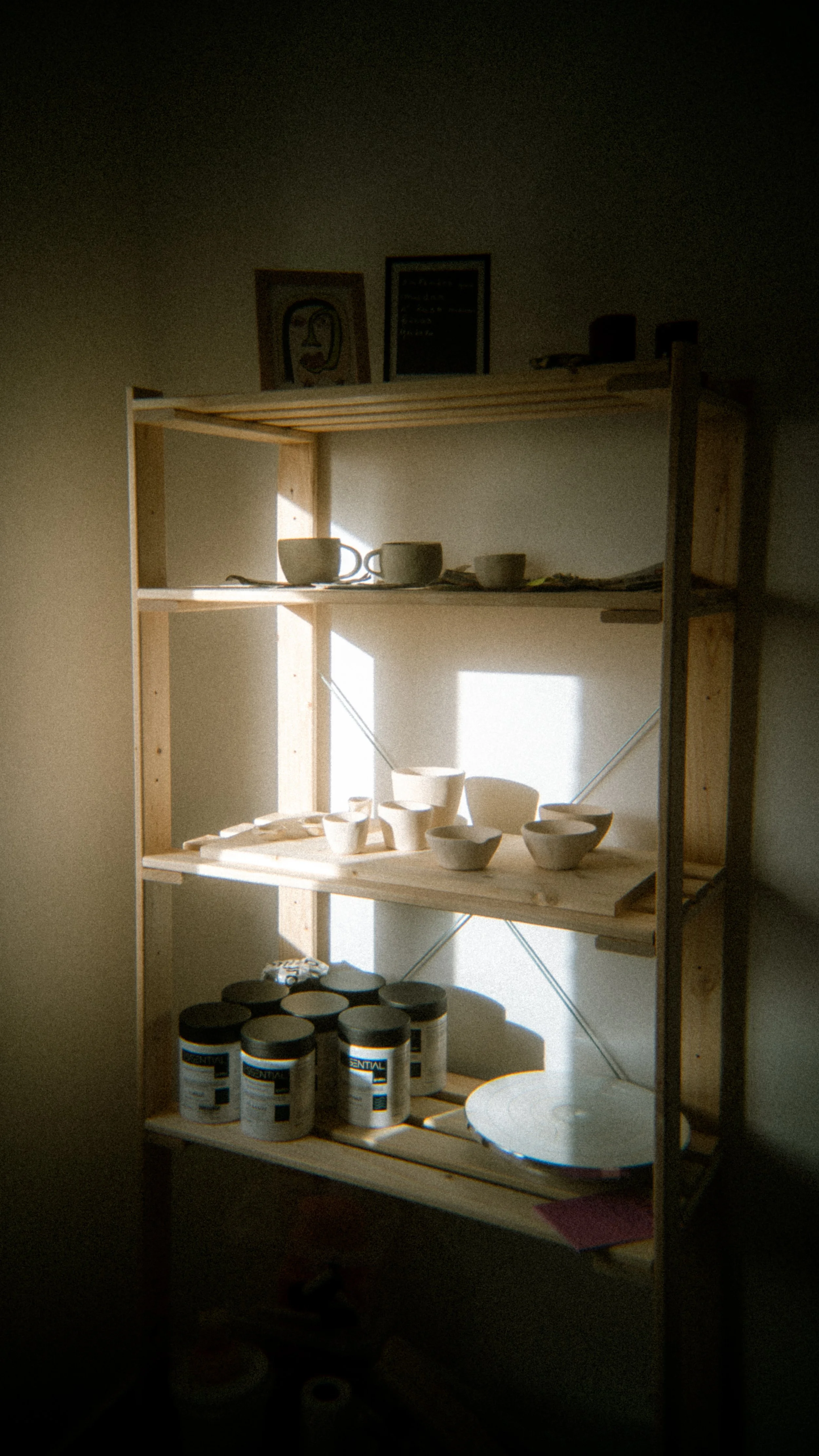 Wooden shelf with pottery mugs, cups and bowls, framed artwork, and containers on the shelves, illuminated by sunlight.