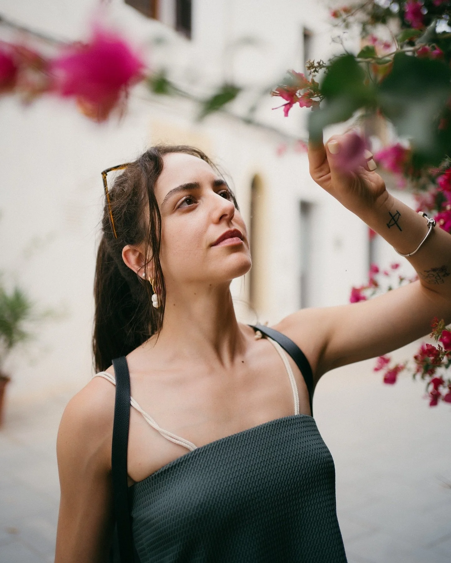 A woman with dark hair, wearing a sleeveless top and sunglasses on her head, is reaching out to touch pink flowers on a plant.