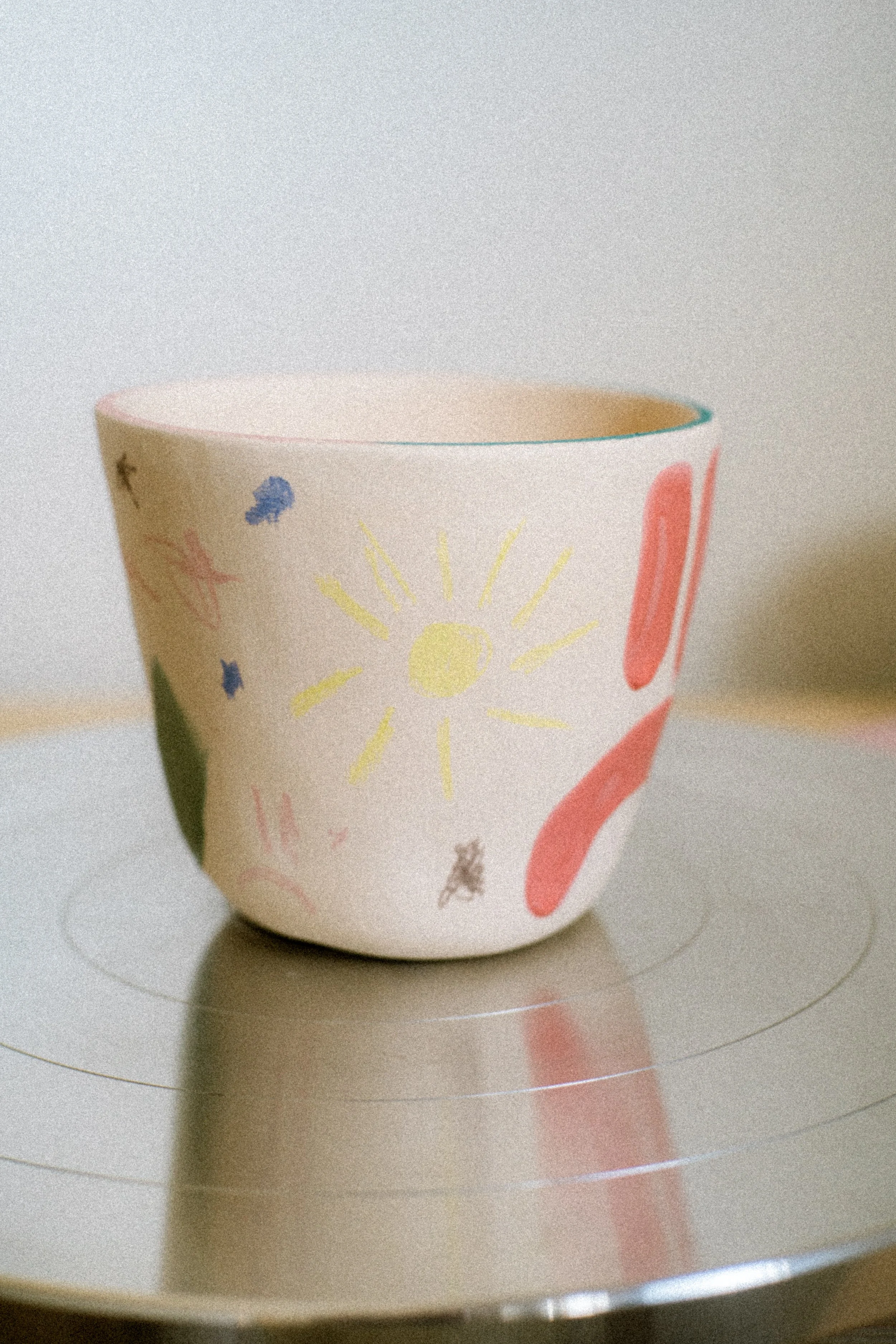 A ceramic bowl with colorful, childlike drawings of a sun, clouds, and abstract shapes, sitting on a reflective potter's wheel.