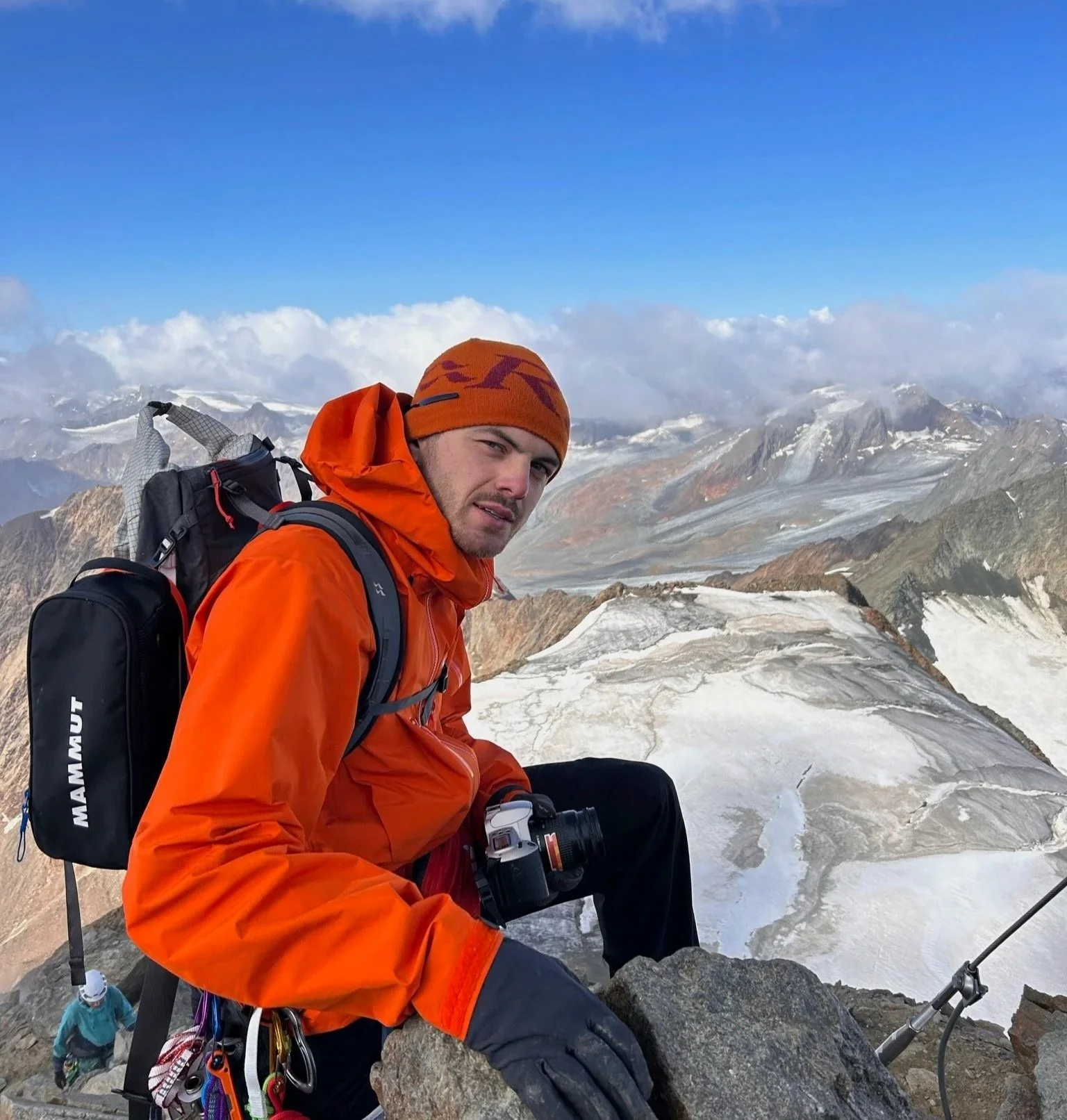 A man in an orange jacket and hat on a mountain peak with snow and rocky terrain, holding a camera, with a climber below him and a mountain landscape in the background.
