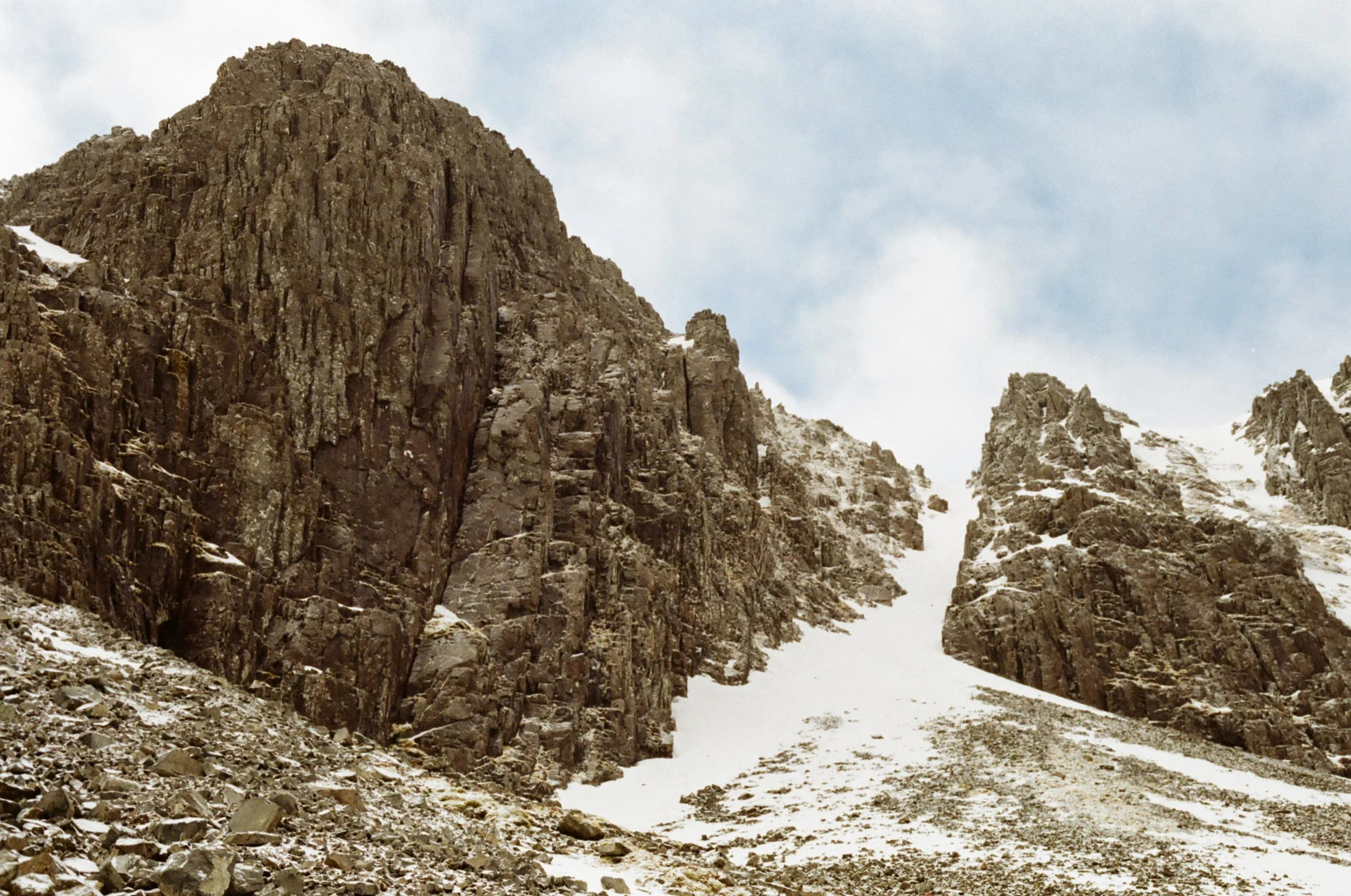 Rocky mountain terrain with large brown rocks and patches of snow, under a partly cloudy sky.