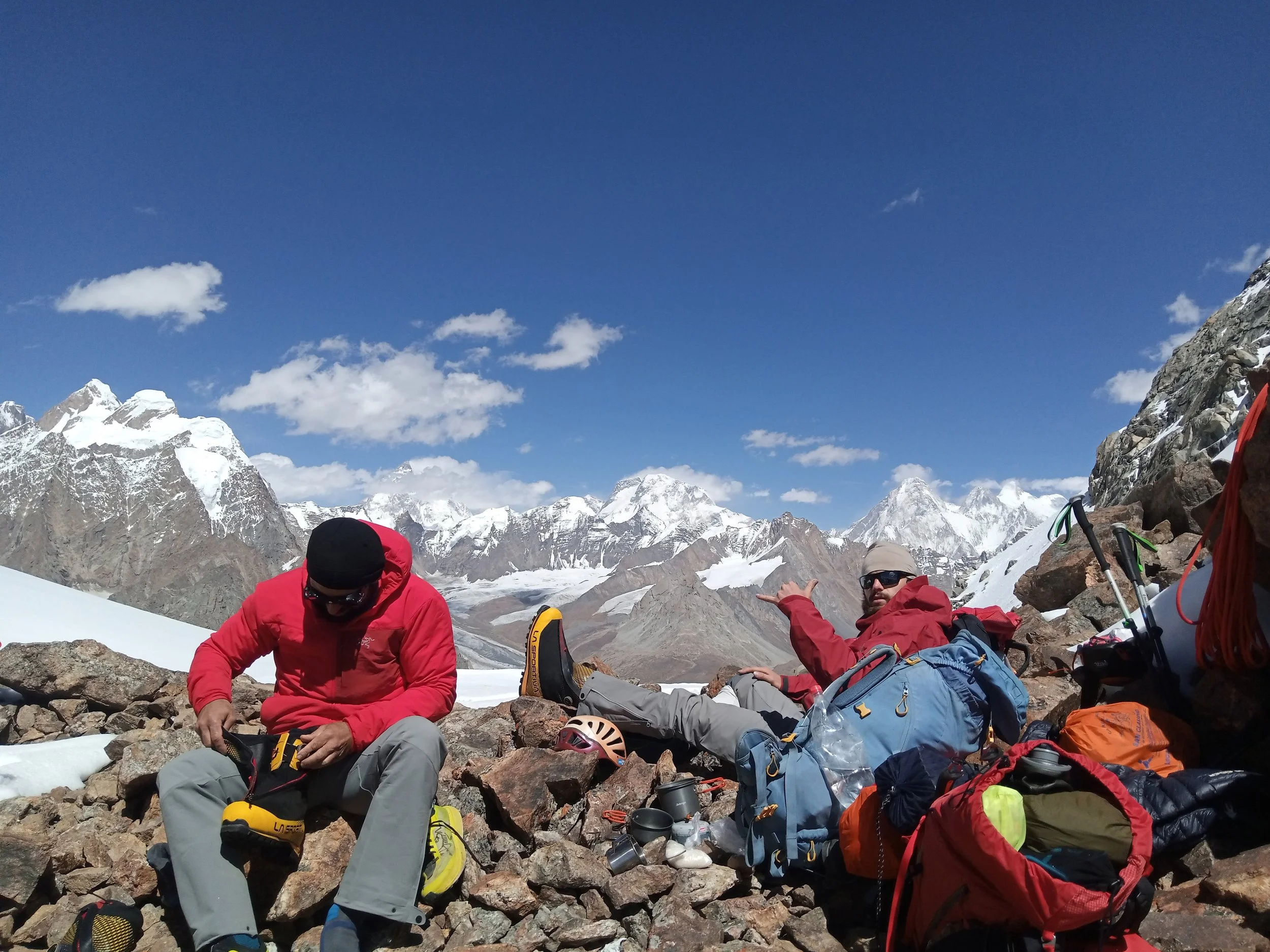 Two mountaineers resting on rocky terrain with snow-covered mountains in the background, one wearing a red jacket and the other in a red jacket, both surrounded by climbing gear.