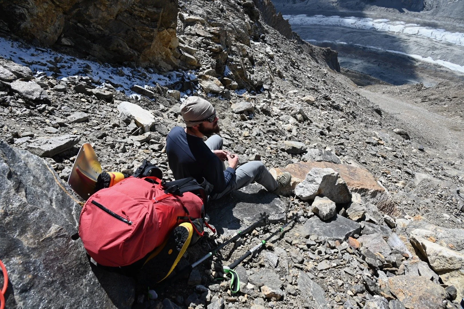 A man wearing outdoor hiking gear, including a beige beanie and sunglasses, sitting on a rocky mountain slope next to his red backpack and trekking poles, with a rugged, snow-capped mountainous landscape in the background.