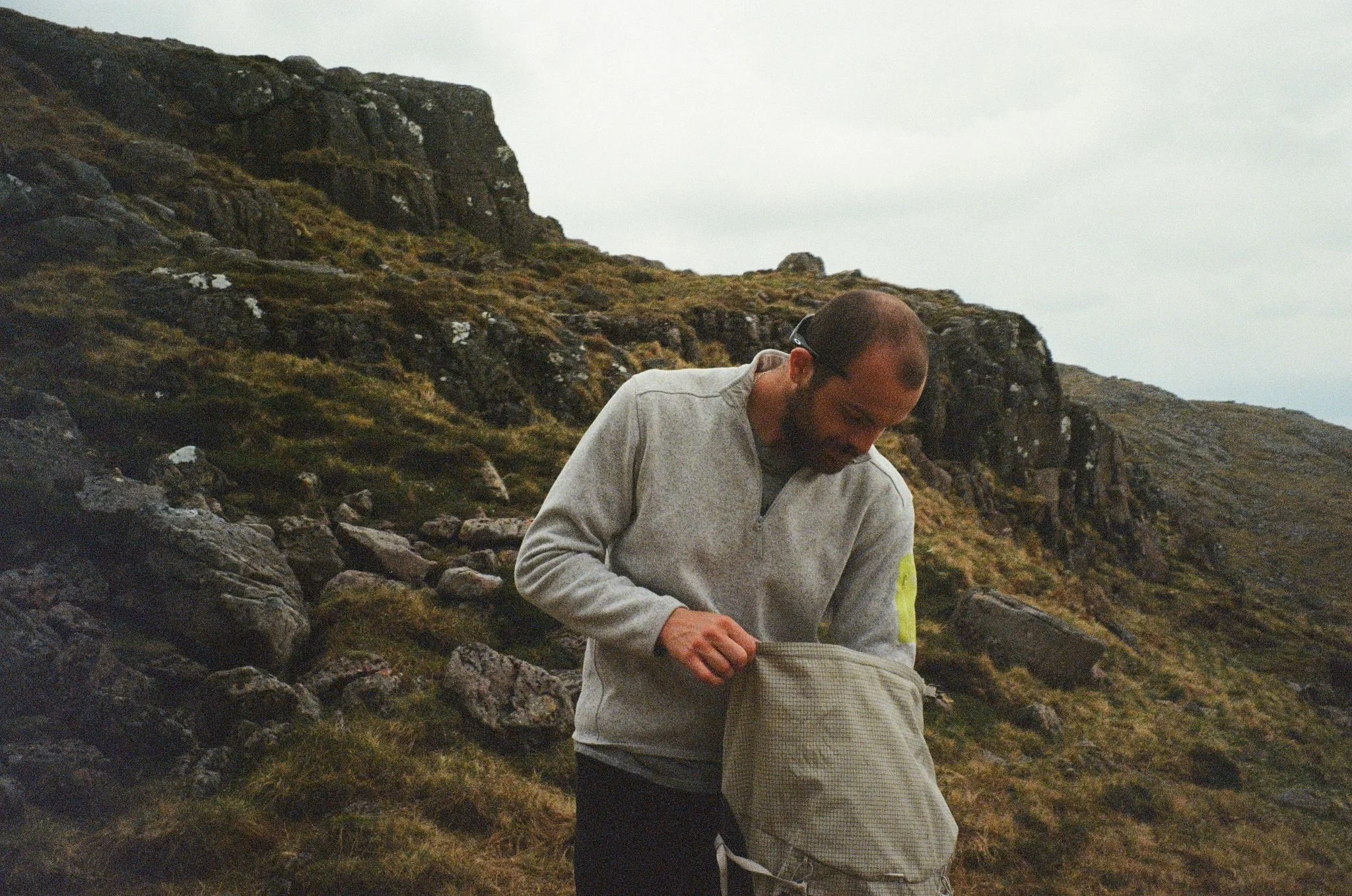 A man with a beard and glasses in a gray sweatshirt looking into a gray backpack outdoors on a rocky, grassy hillside with mountains in the background.