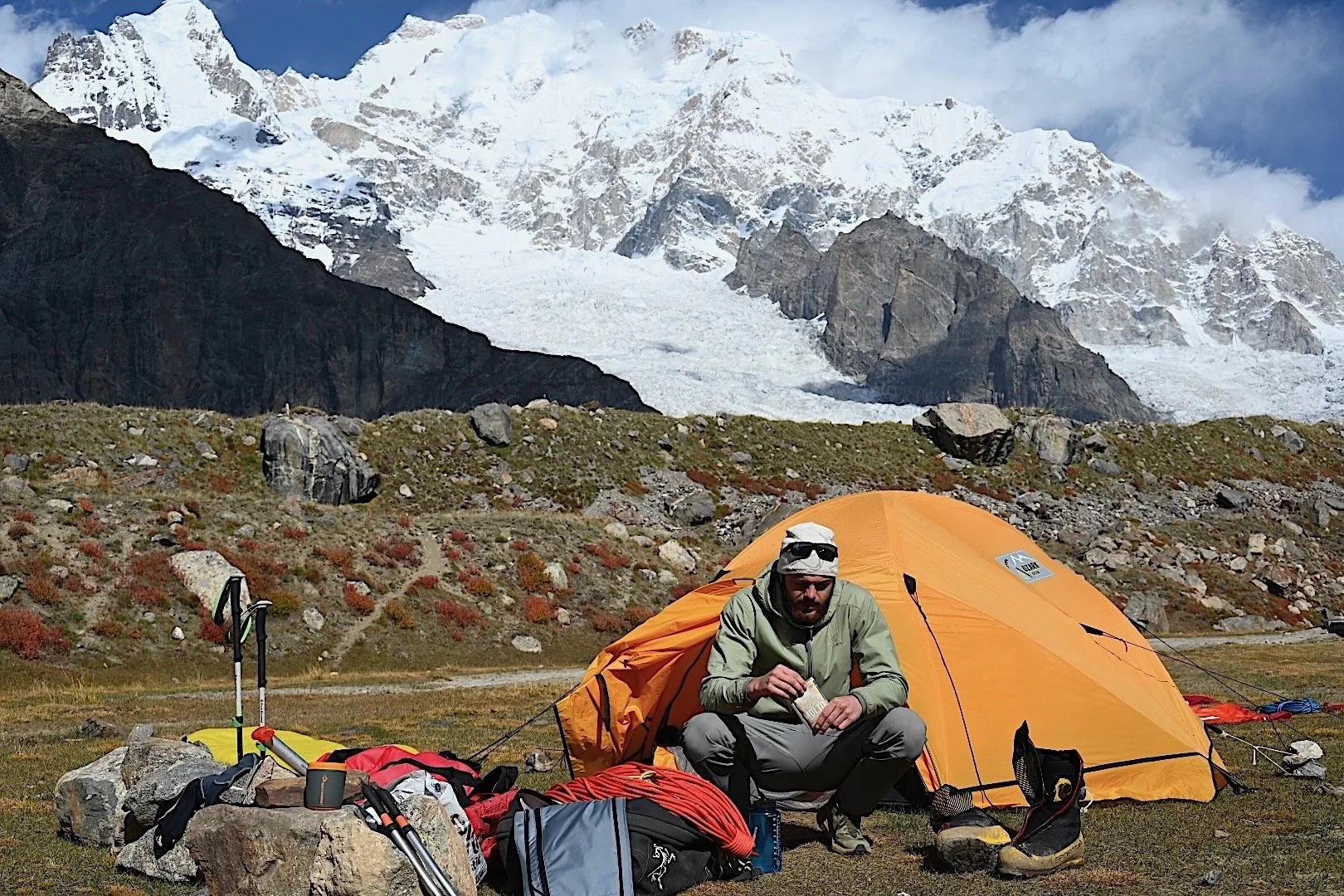 A man camping outdoors with a yellow tent, surrounded by gear such as hiking poles, backpacks, and shoes, in a mountainous area with snow-covered peaks in the background.