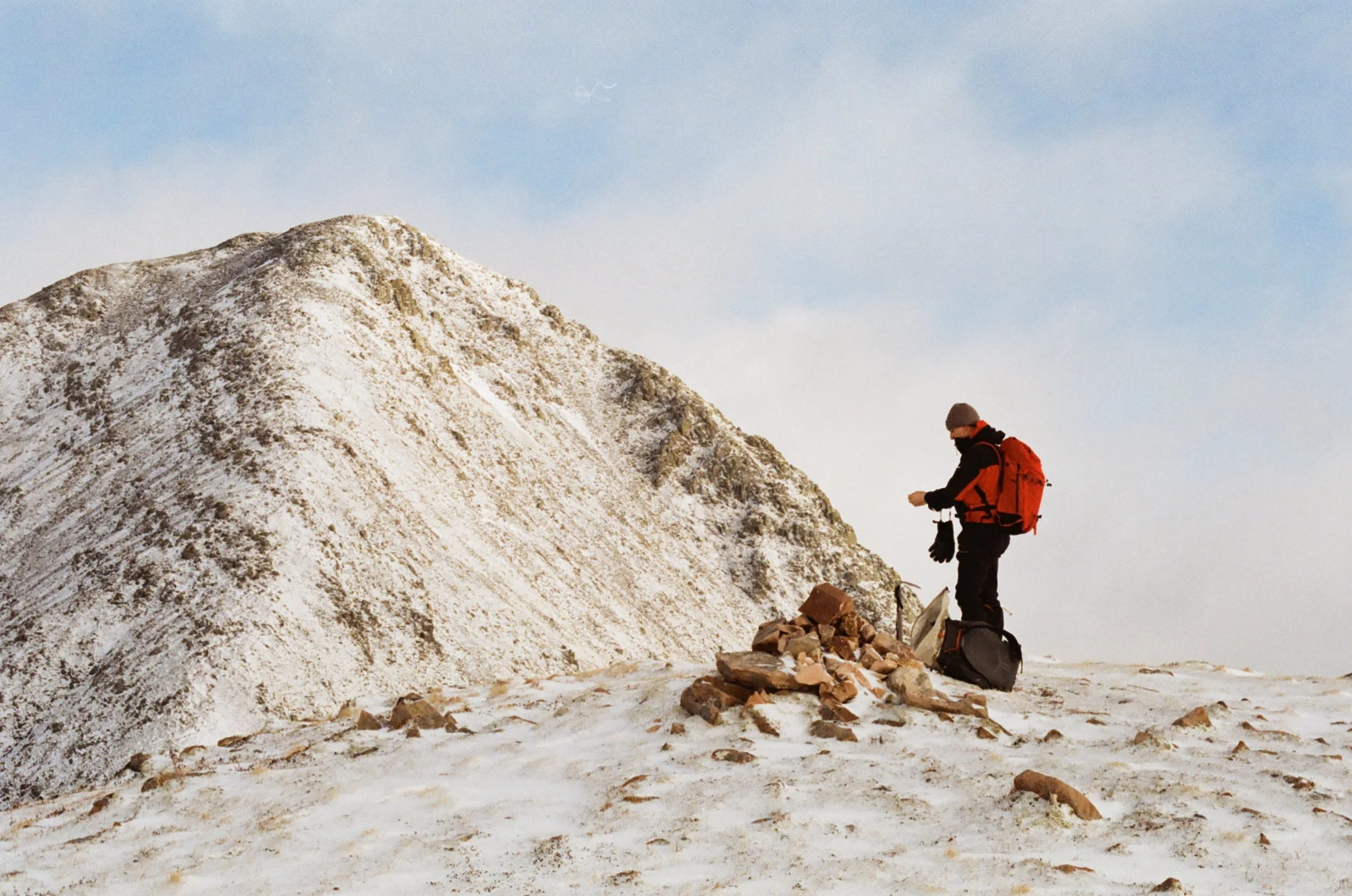 A person dressed in winter gear, including a gray beanie, black jacket, and carrying an orange backpack, stands on snowy ground near a small pile of rocks in a mountainous, snow-covered landscape under a partly cloudy sky.