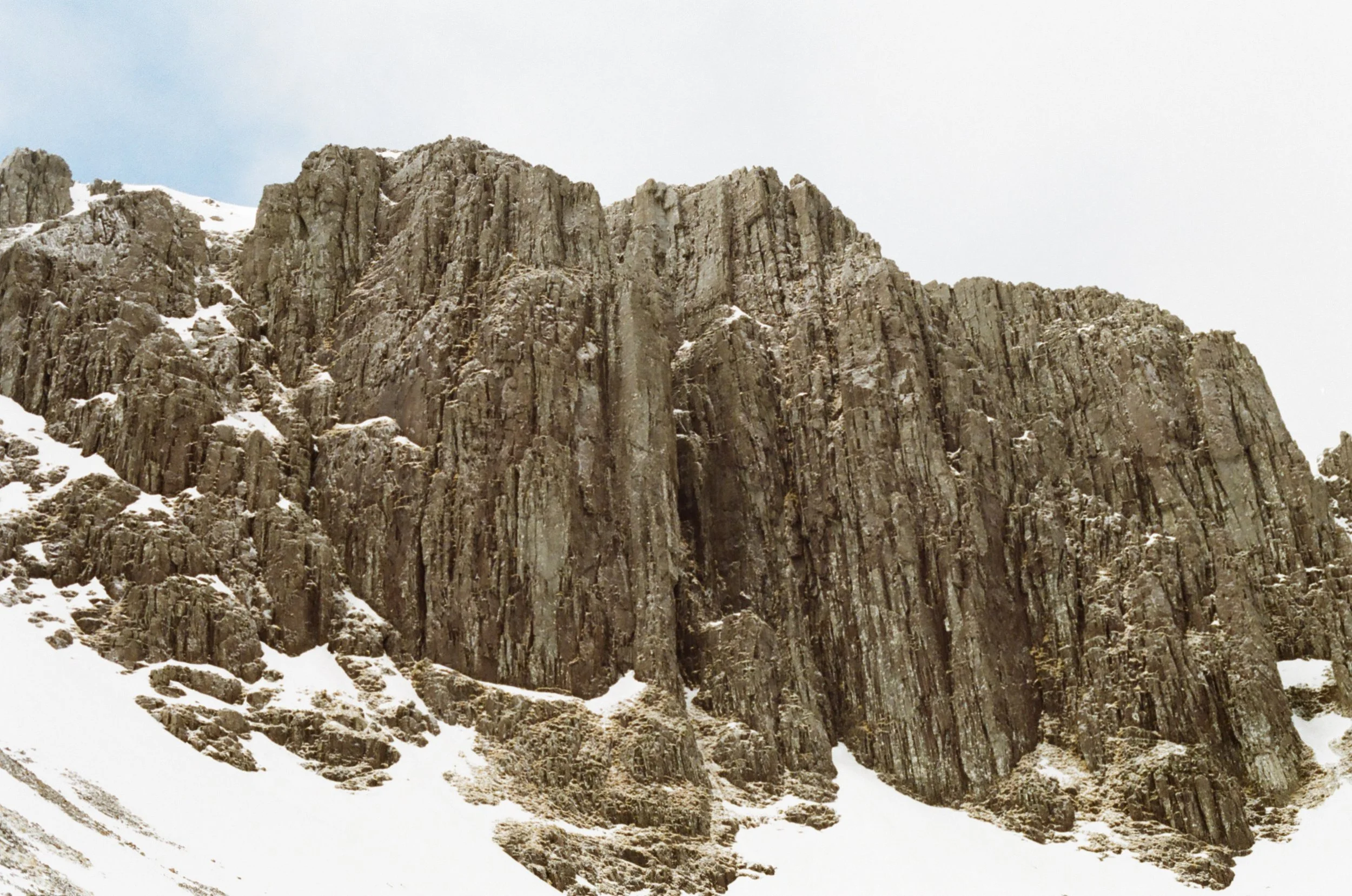 Snow-covered mountain with steep, rugged rocky cliffs under a cloudy sky.