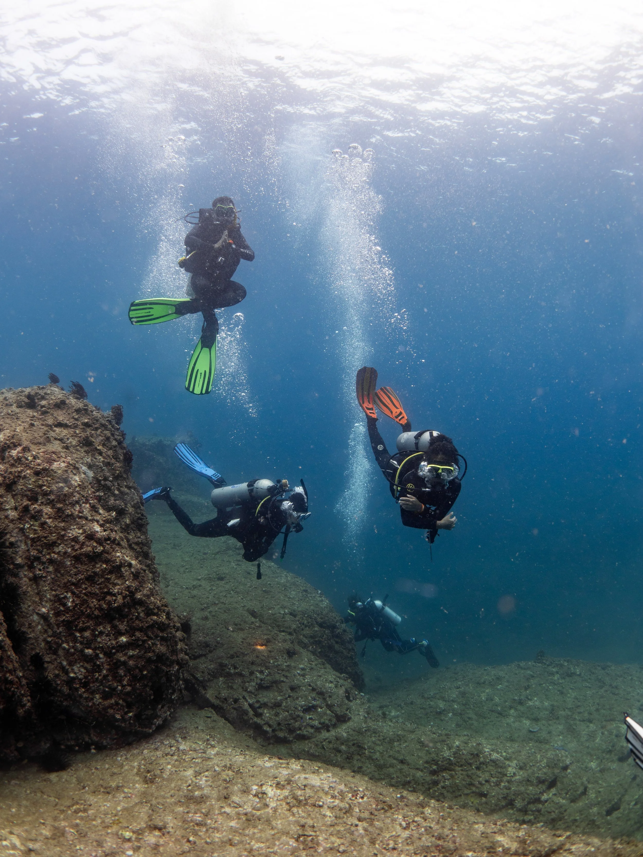 Three scuba divers exploring underwater near rocks and coral with bubbles rising to the surface.