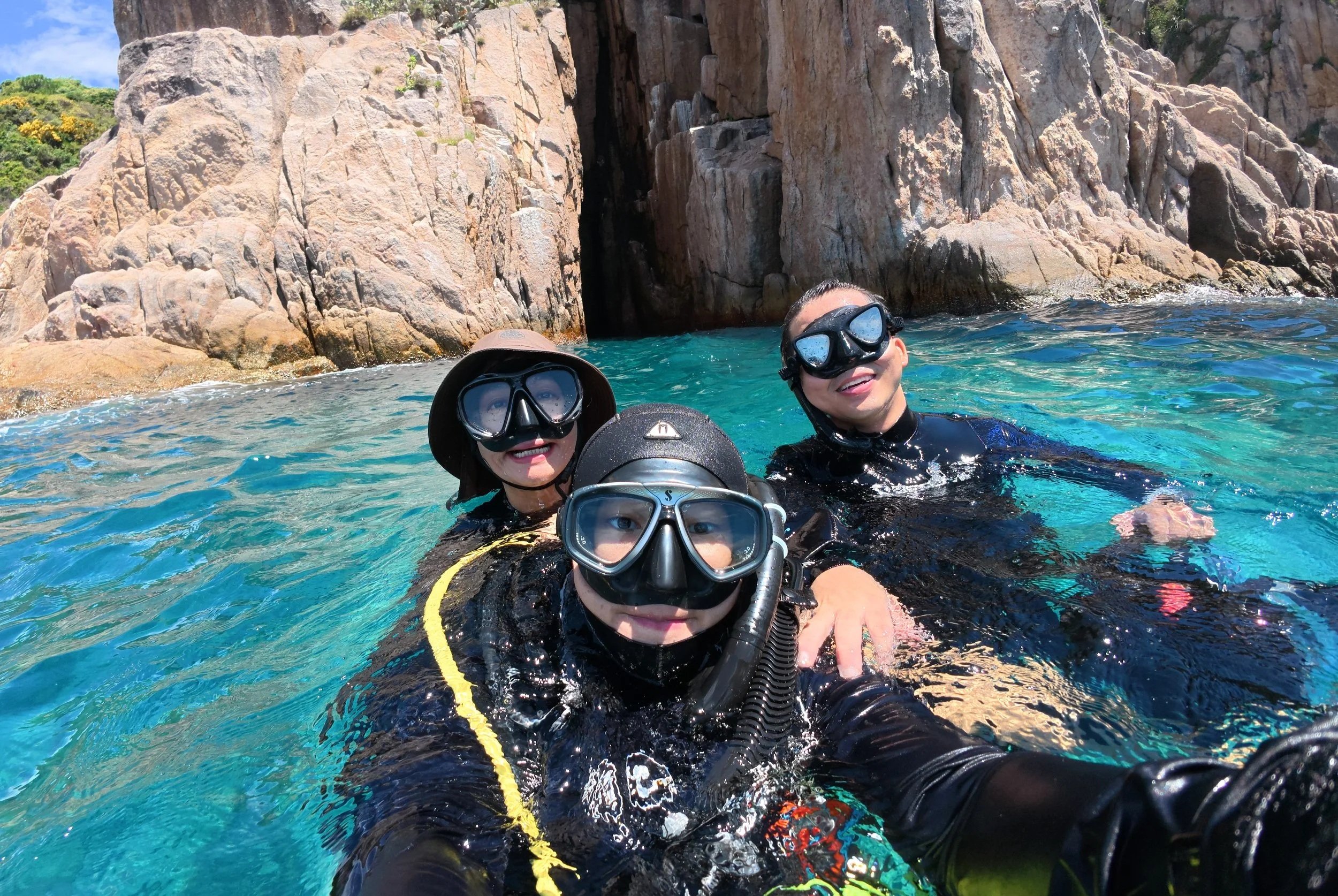 Three people in scuba gear taking a selfie in clear blue water near rocky cliffs.