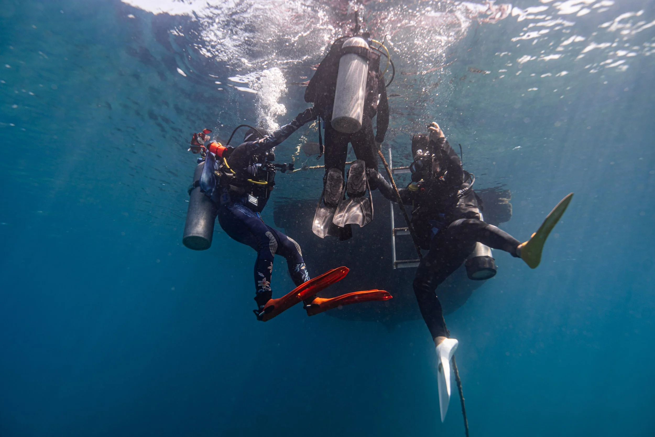Two scuba divers assisting a person with a scooter underwater, with the diver on the left holding the scooter's handlebars and the diver on the right supporting from below.