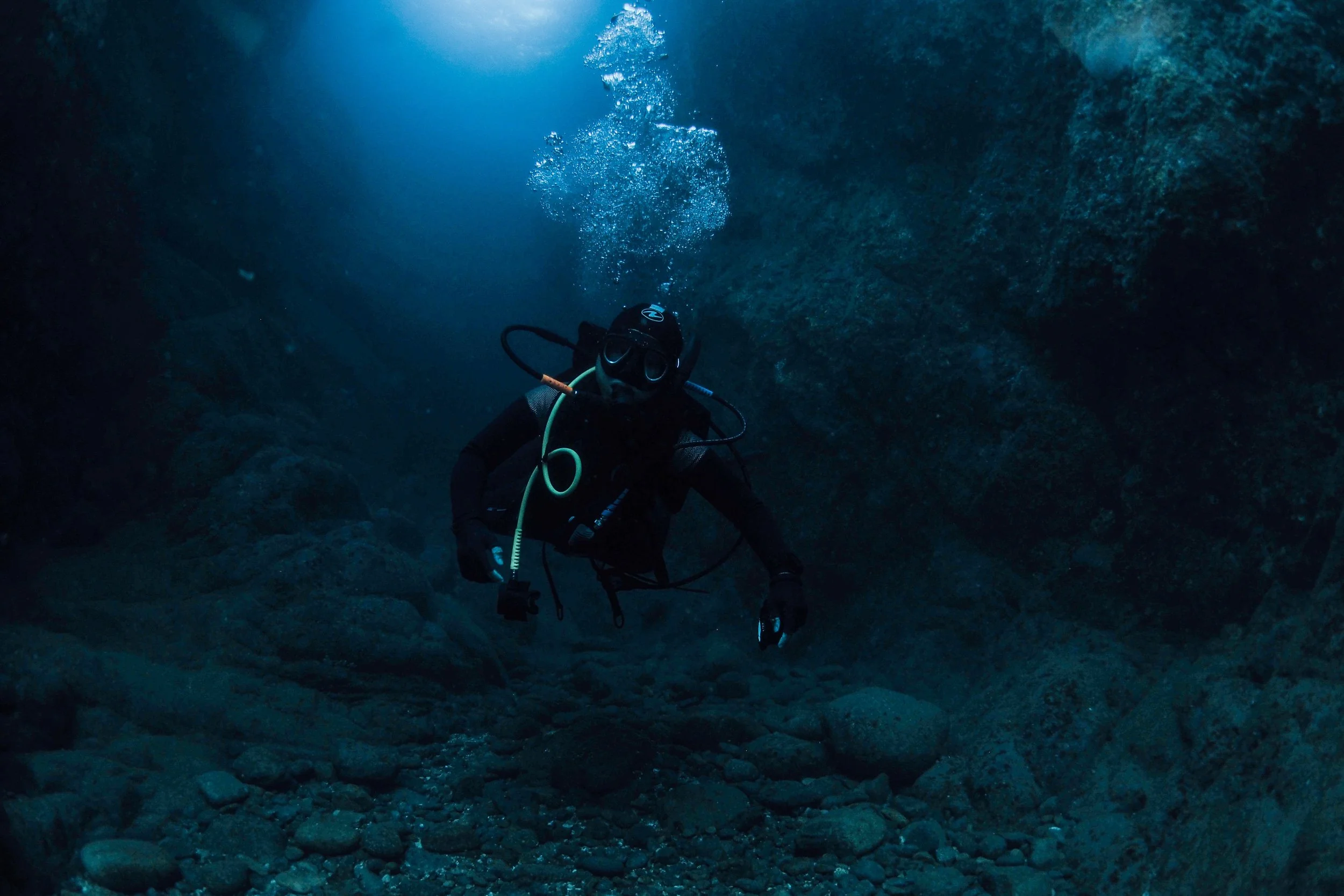 A scuba diver exploring an underwater cave or crevice with rocky walls and gravel on the bottom, beams of light filtering from above.
