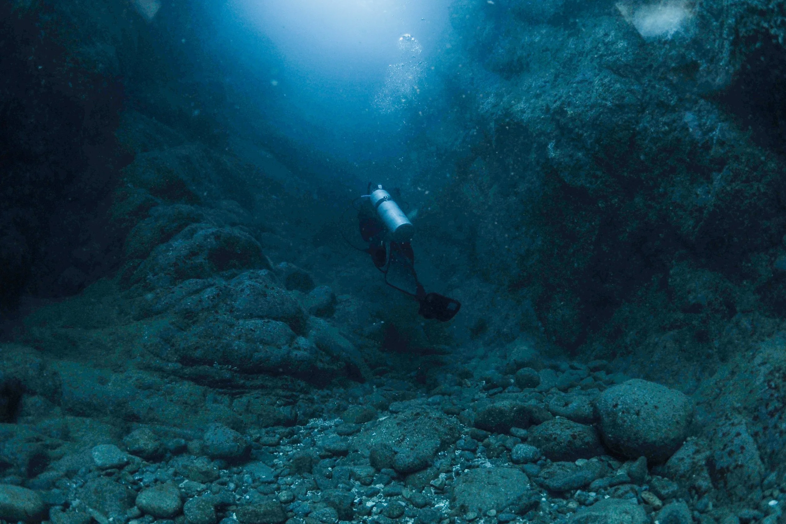 An underwater scene with a scuba diver swimming through a rocky canyon.