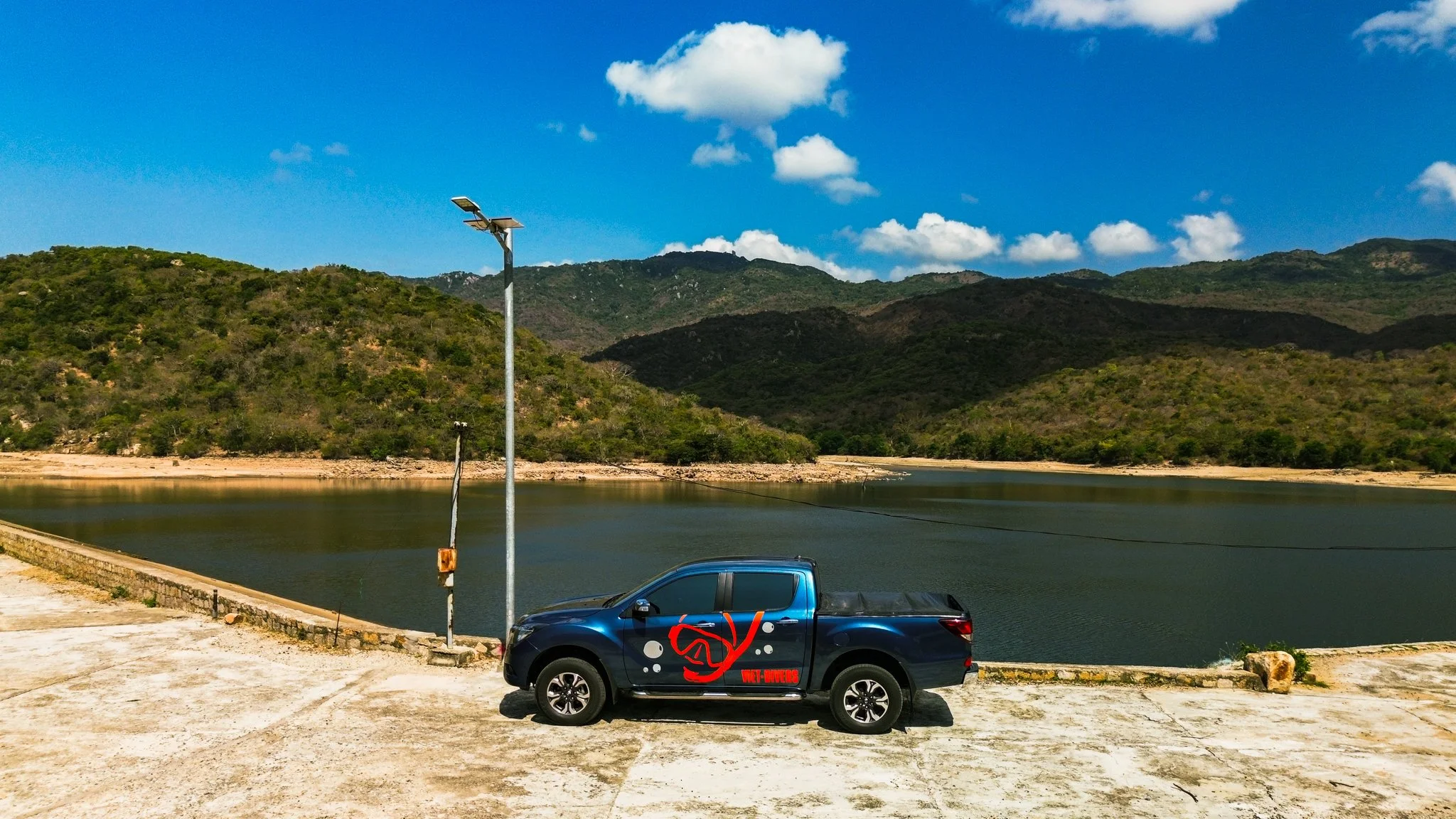 Blue pickup truck parked near a body of water with hills in the background under a sunny sky.