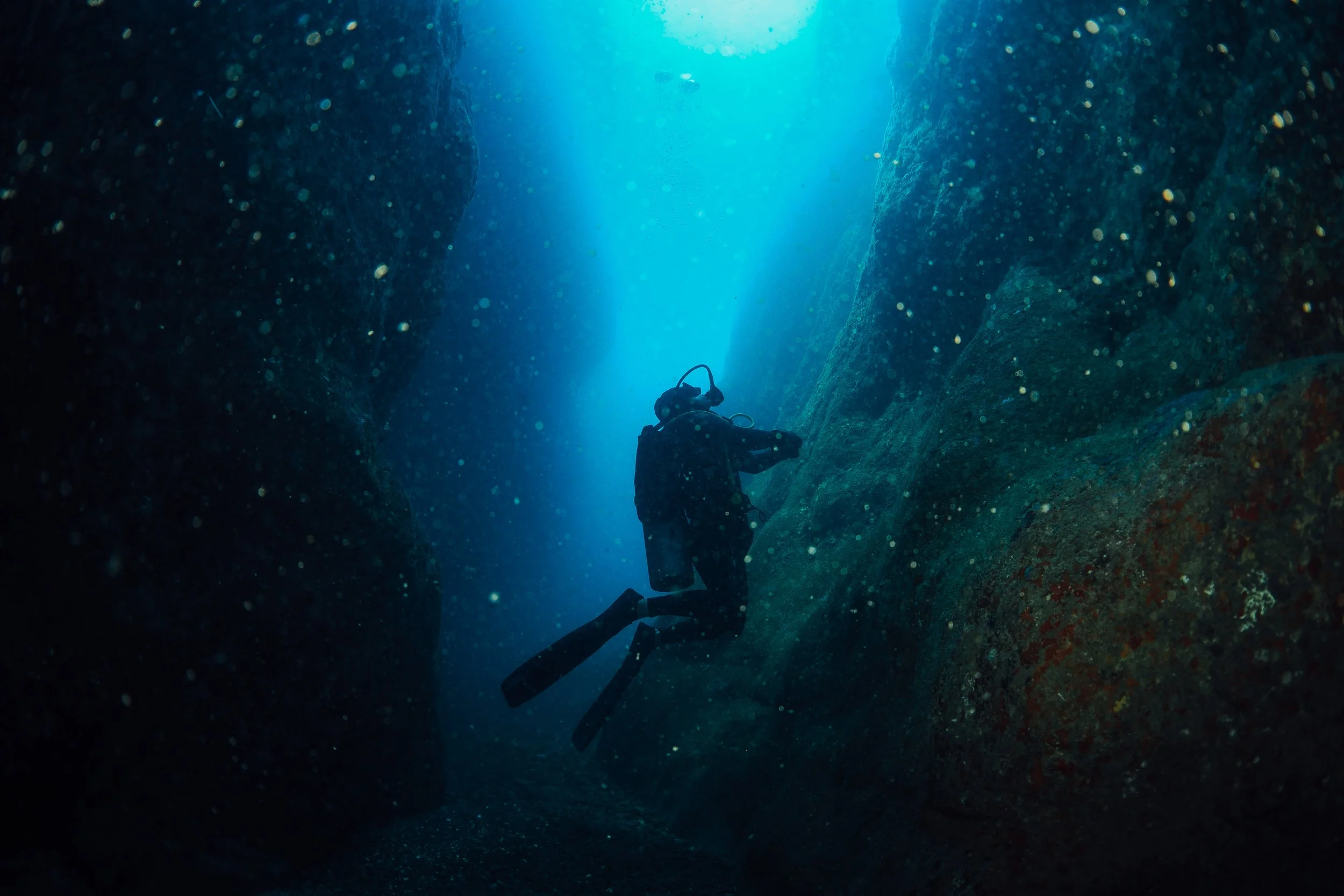A scuba diver swimming through a narrow underwater canyon with light shining from above.