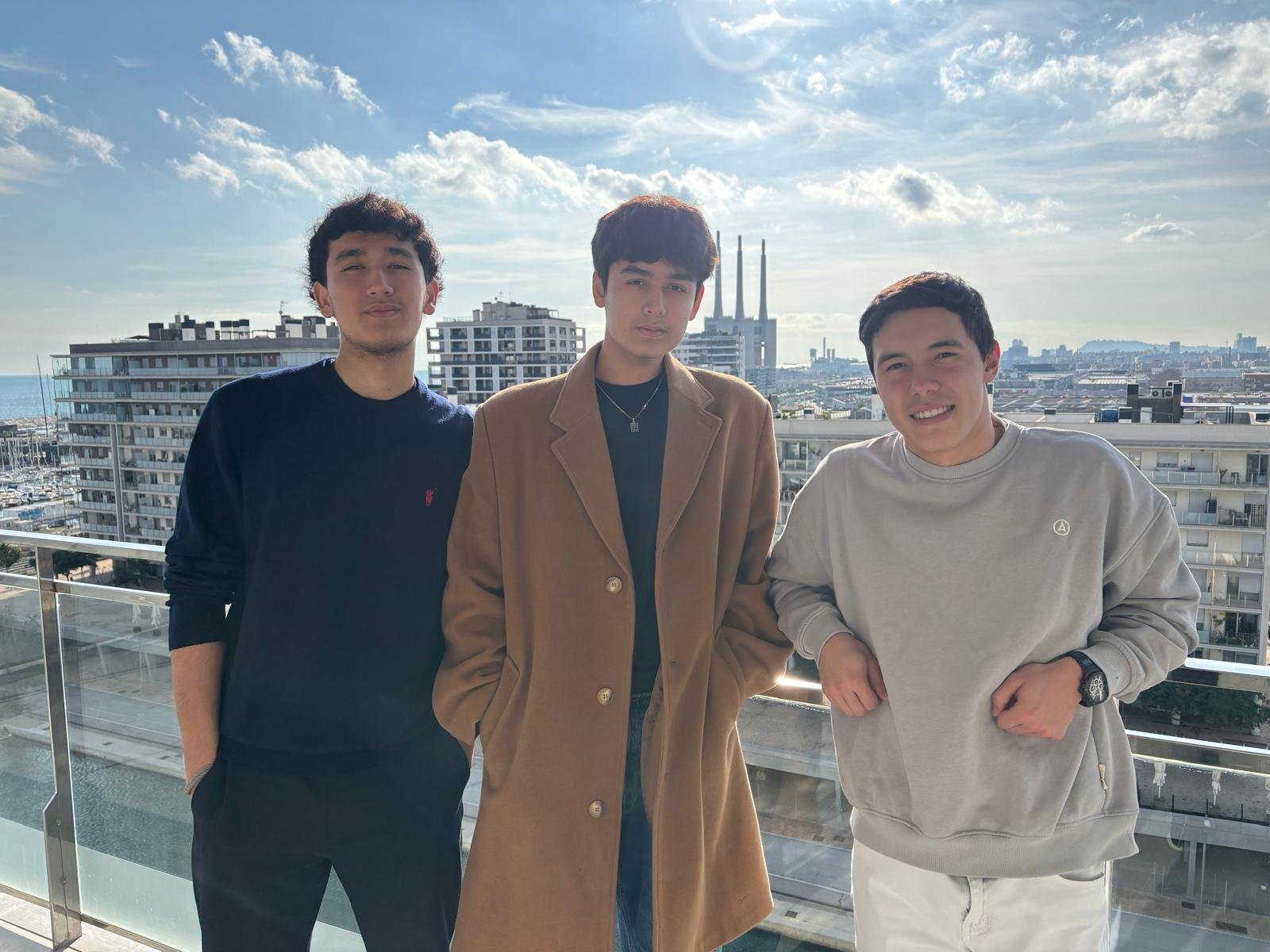 Three young men standing on a balcony with a cityscape and water in the background, under a partly cloudy sky.
