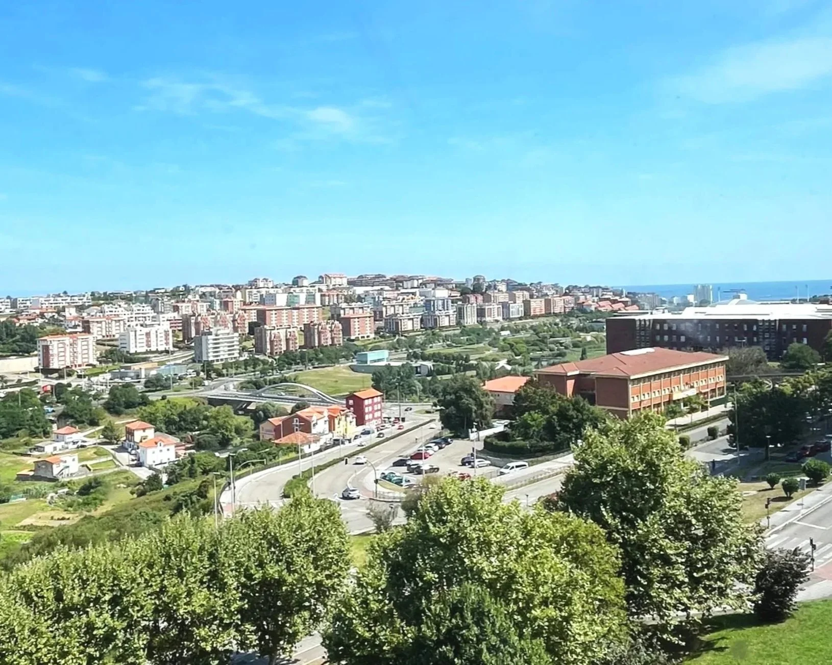 View of a city with high-rise buildings and apartments on hilly terrain, green trees in the foreground, and a bright blue sky.