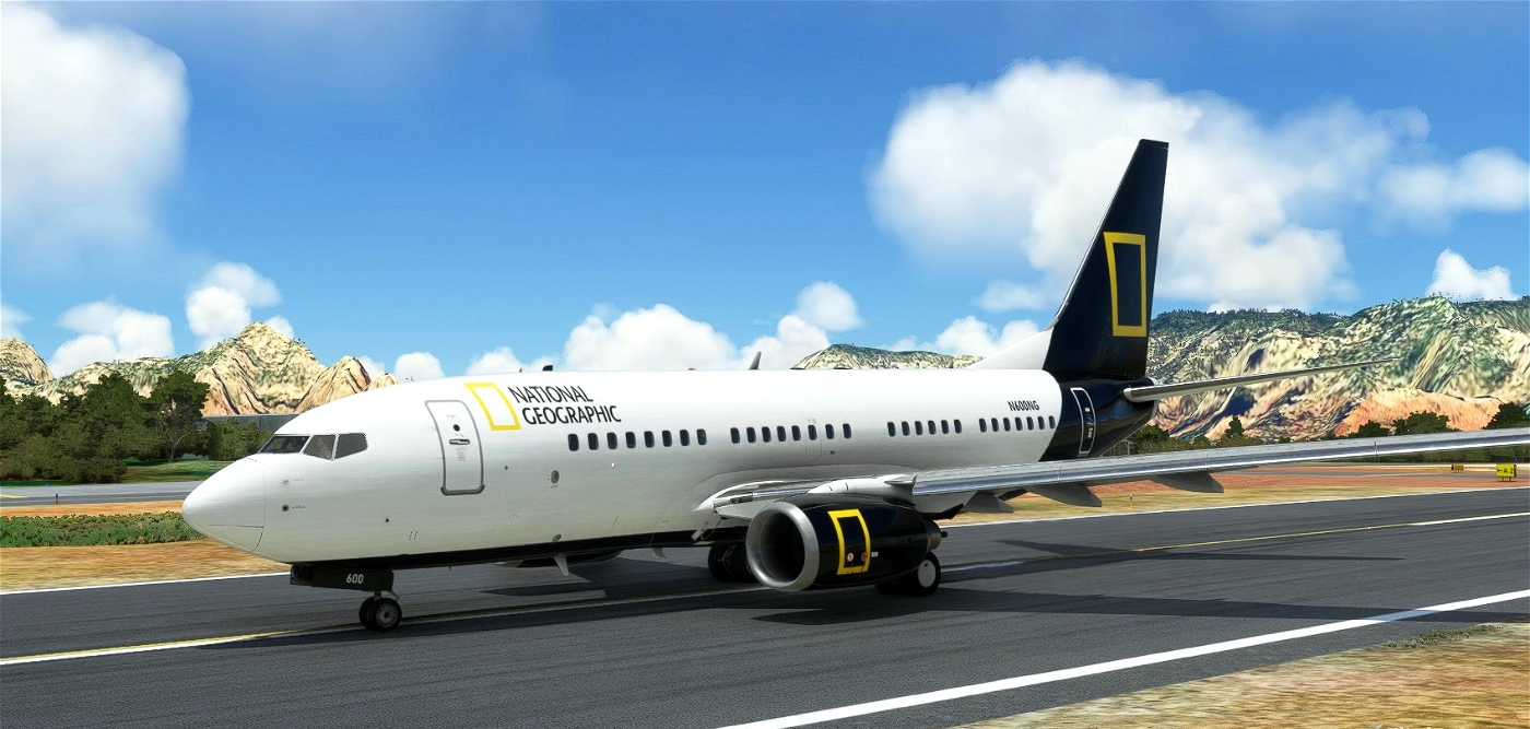 Commercial airplane with National Geographic logo on the side and tail, parked on runway with mountains and blue sky in background.