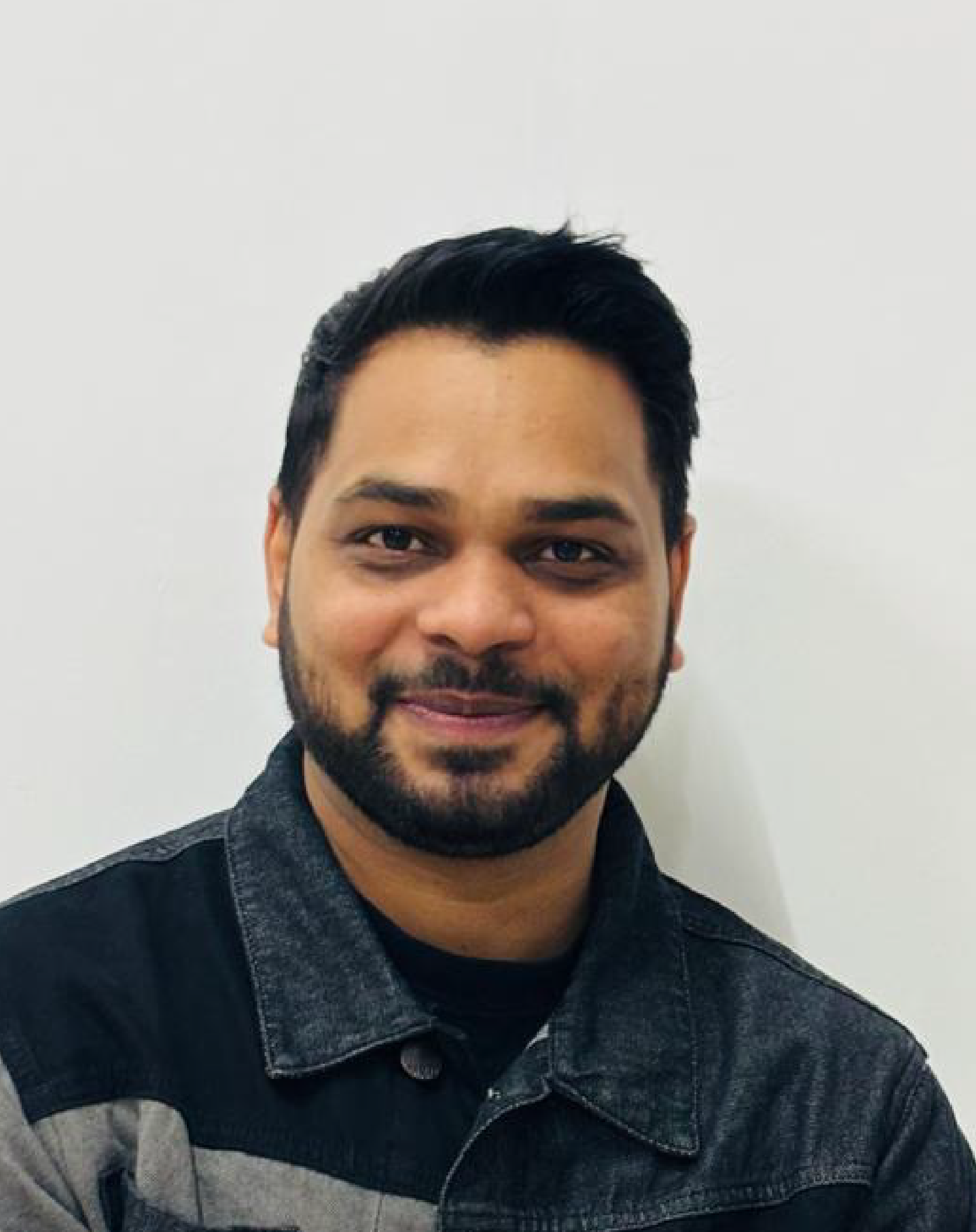Close-up portrait of a young man with dark hair, beard, and mustache, wearing a black and gray denim jacket, smiling against a plain white background.