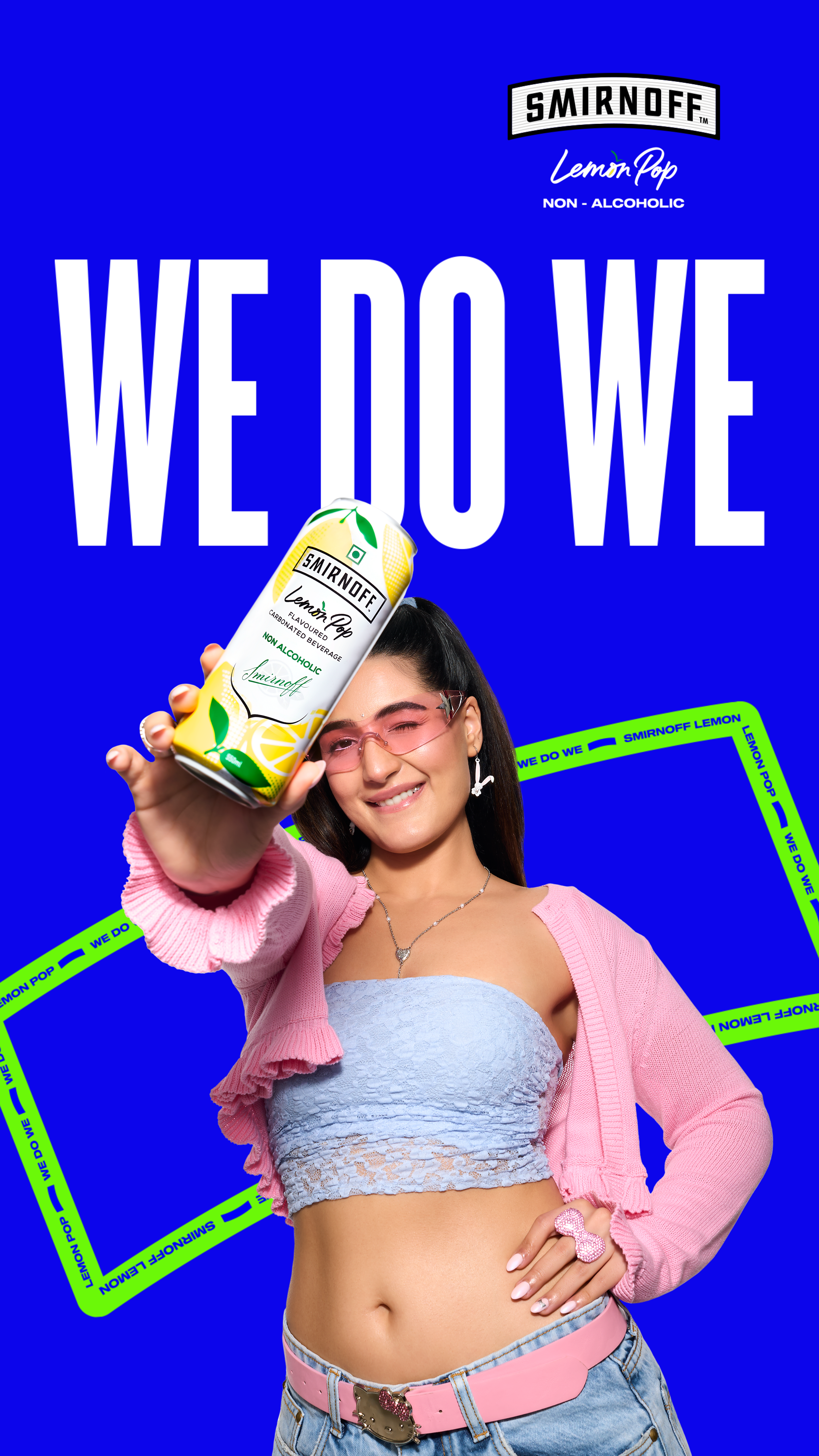 A woman in a pink cardigan and light blue lace crop top holding a can of Smirnoff Lemon Pop non-alcoholic beverage, smiling, with a vibrant blue background and green decorative tape that reads "We Do We" and "Smirnoff Lemon Pop".