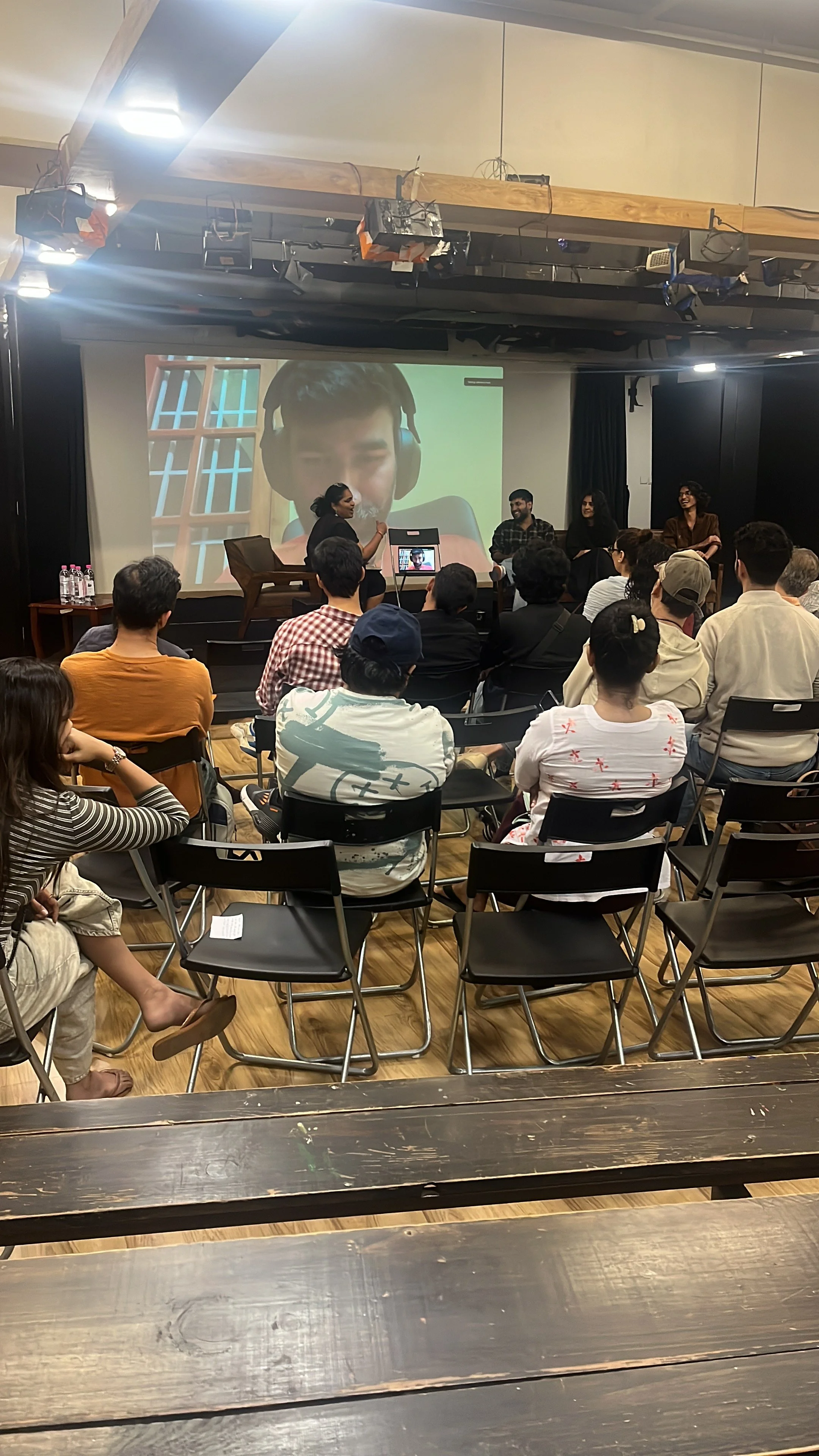 A group of people attending a presentation or panel discussion in an auditorium. There are five panelists seated at the front, a woman speaking, and a large screen behind them displaying a young person's face wearing headphones. The audience is seated in black chairs, and the room has a wooden floor and black curtains on the sides.