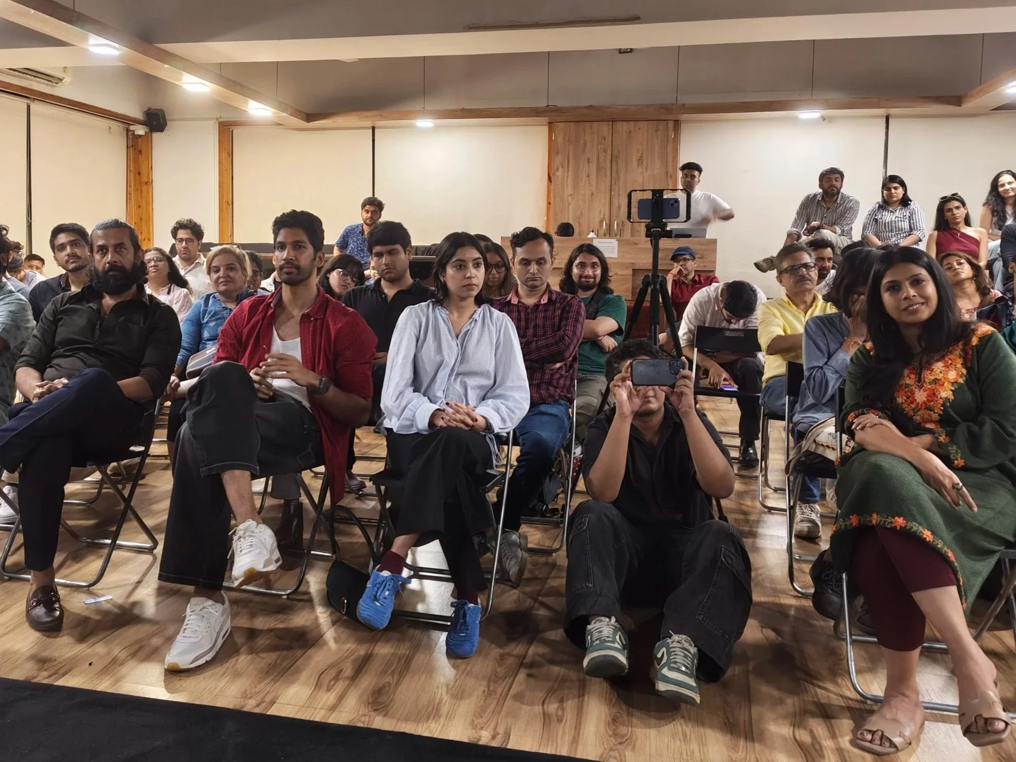Audience seated indoors, some taking notes or using phones, with a person standing near a camera tripod at the back.