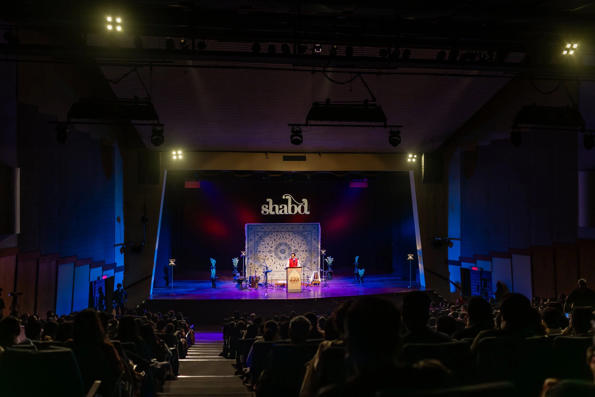 A stage in an auditorium with a performer standing at a podium, decorated with a backdrop and floral arrangements, in front of an audience seated in the dark. The setting appears to be for a cultural or musical event at Shabd.