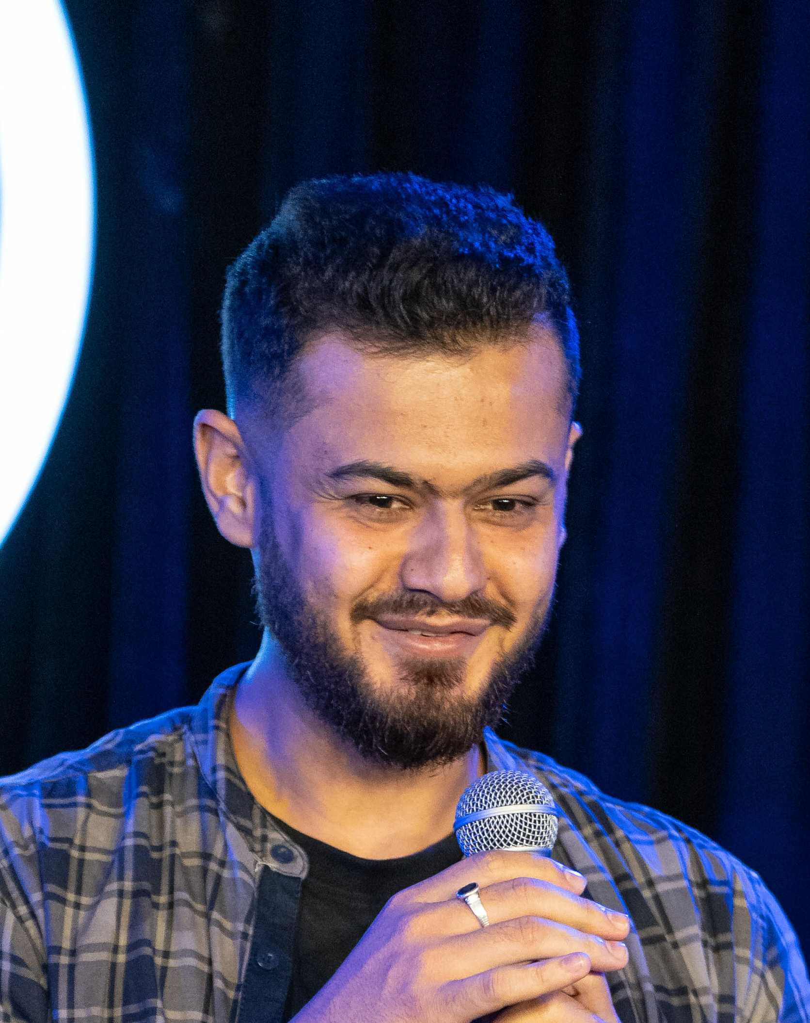 A man with short dark hair and a beard, holding a microphone and smiling, standing in front of a dark background.