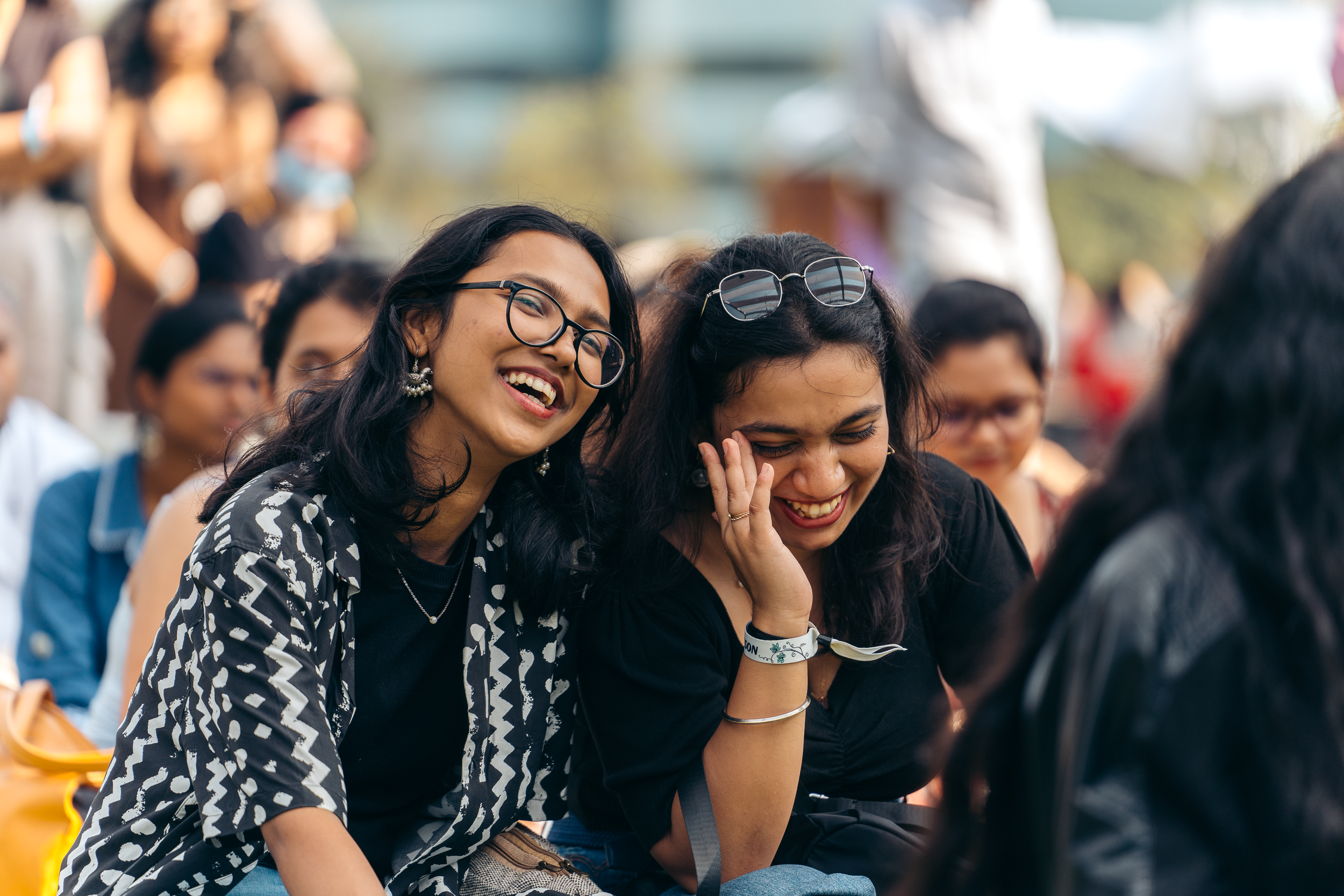 Two young women laughing and smiling at an outdoor event, with one wearing glasses and patterned shirt, and the other with sunglasses on her head and wearing a black top, surrounded by other people in the background.