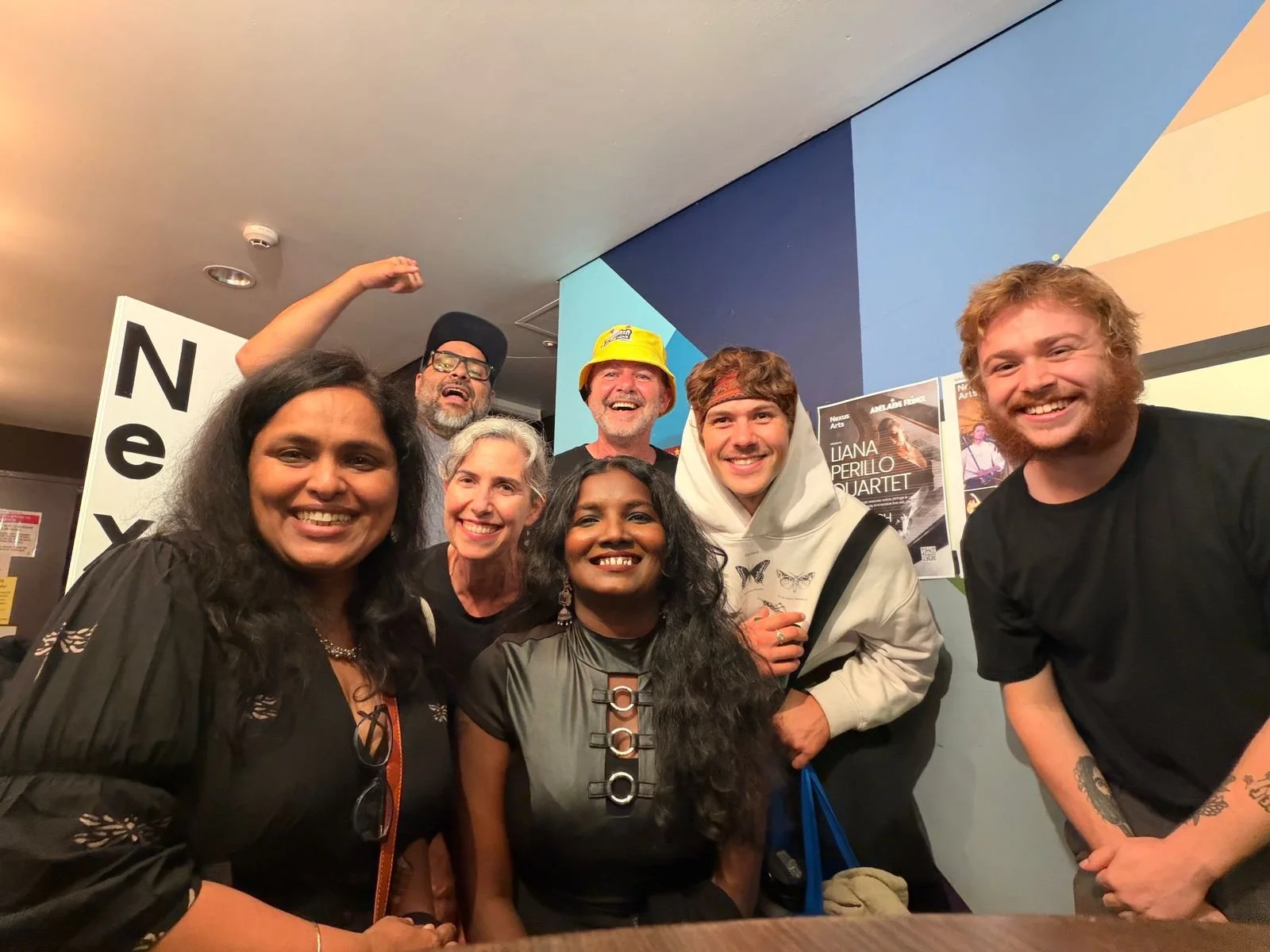 Group of seven diverse people smiling and posing for a photo indoors, some making playful gestures, including a woman with long black hair, in front of a blue wall with posters and signs.