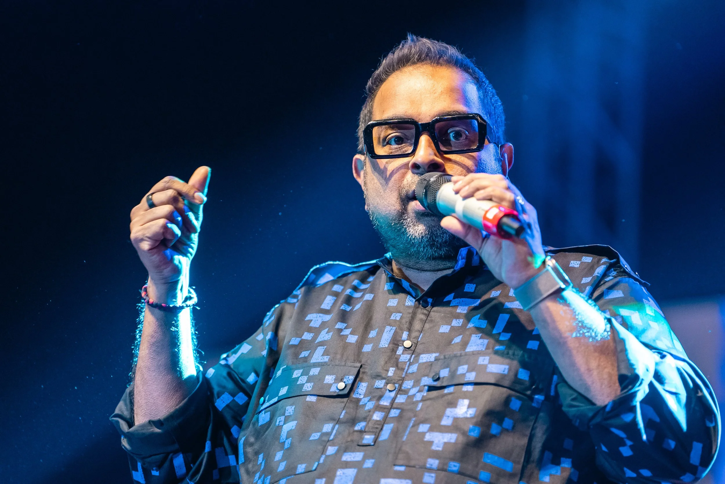 A man with dark hair and glasses singing into a microphone on stage, wearing a patterned shirt under blue lighting.