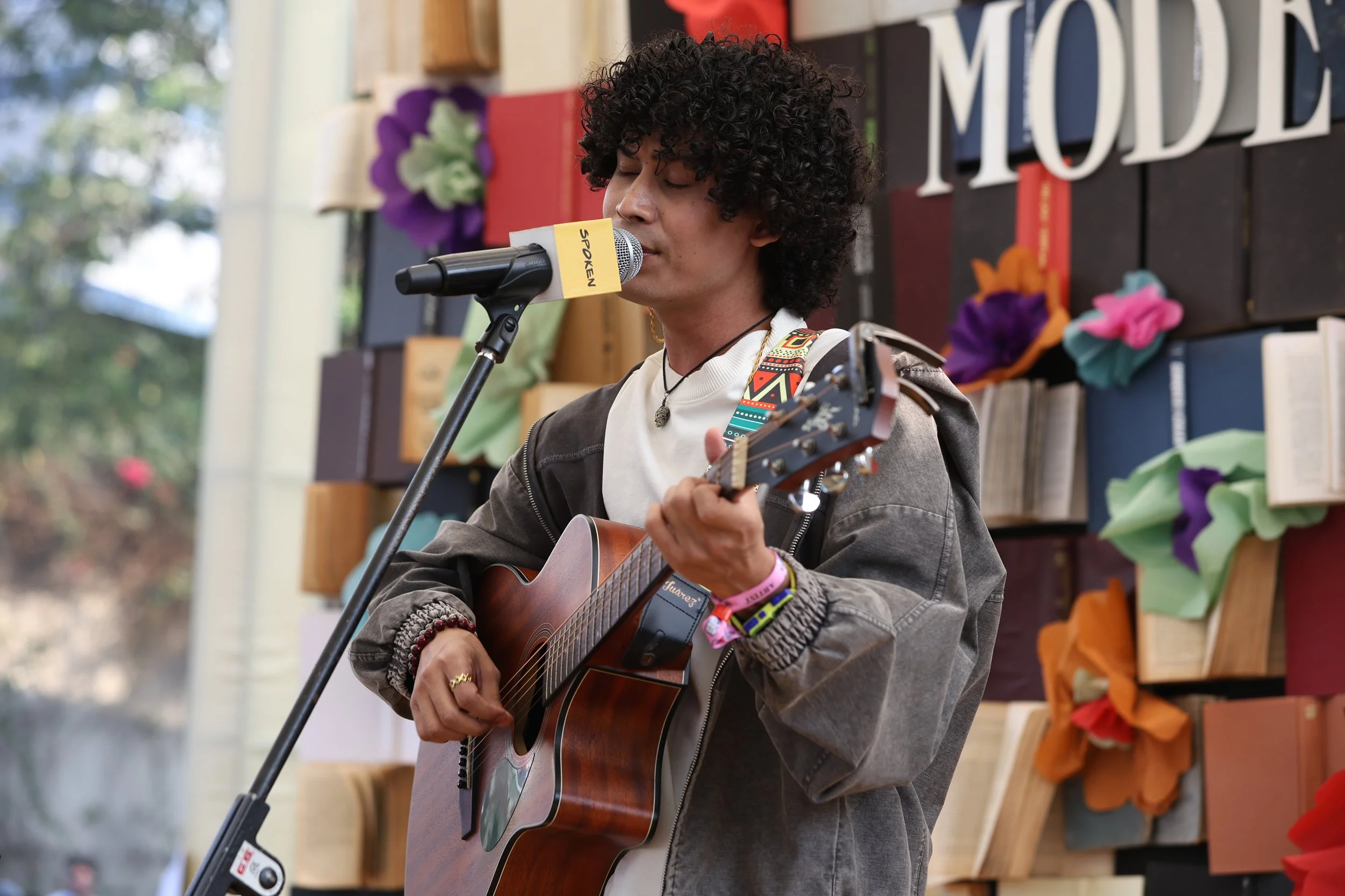 A person (Maan Panu) with curly hair is playing an acoustic guitar and singing into a microphone. They are standing indoors, with a colorful backdrop including books and paper flowers.