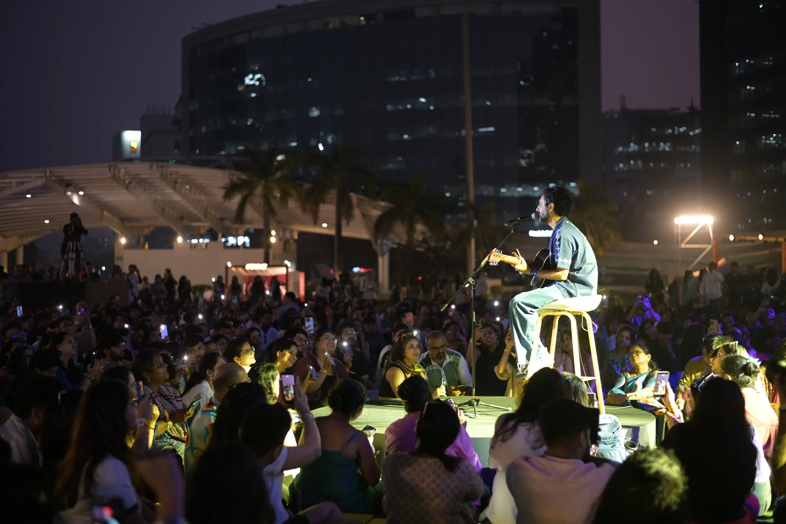 A man performs on an elevated platform playing guitar and singing into a microphone during an outdoor concert at night. The audience is sitting and watching attentively, capturing the moment on their phones. Tall modern buildings are visible in the background under a dark sky.