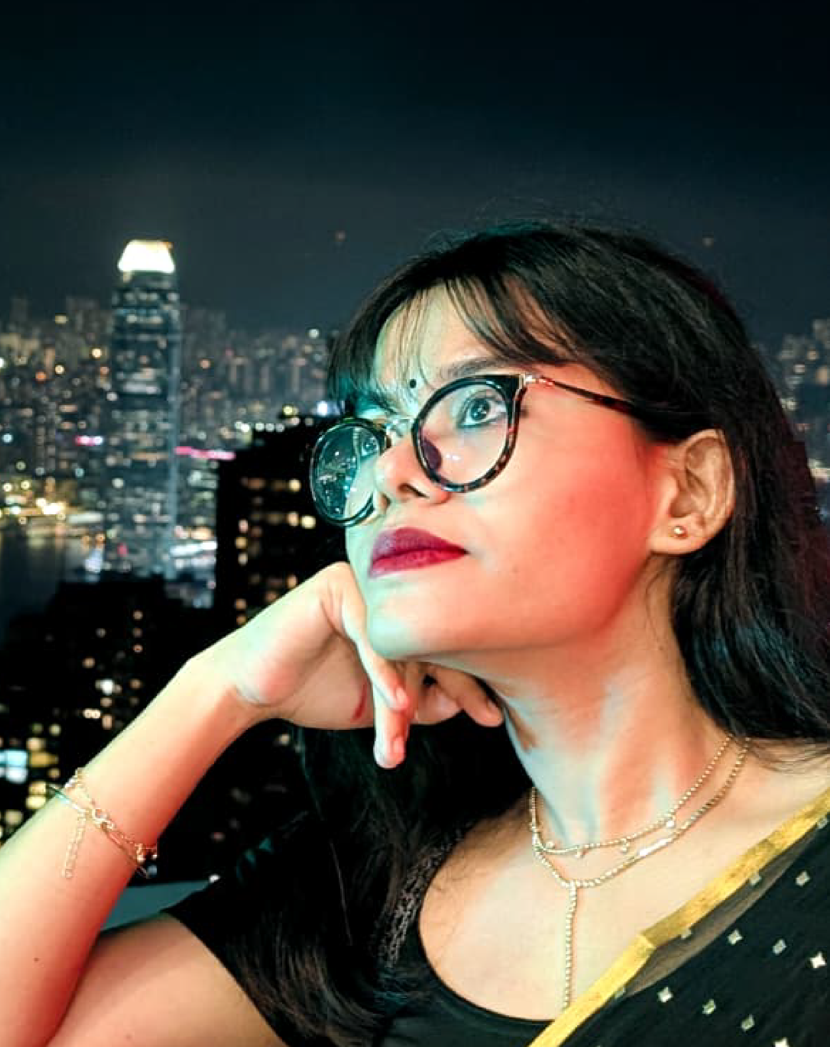 Young woman with glasses and jewelry looking thoughtfully at the night city skyline from a high-rise building.